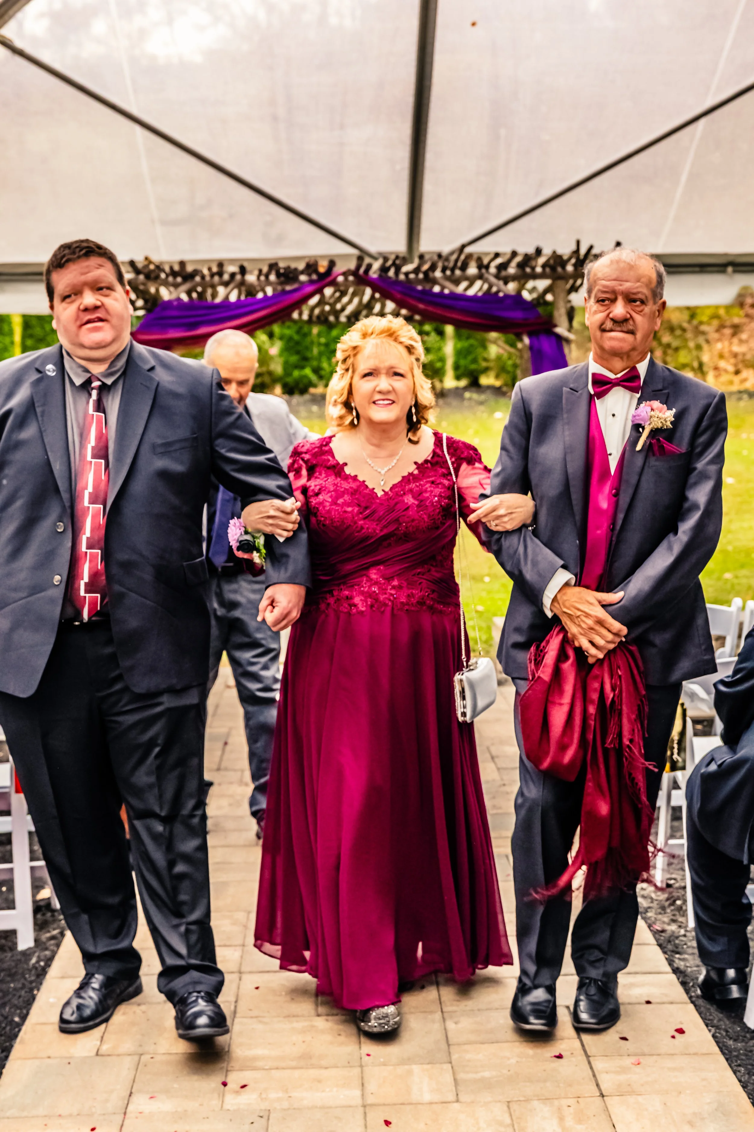 A woman in a burgundy dress walking down the aisle towards a wedding ceremony, flanked by two men in suits, under a decorated canopy.