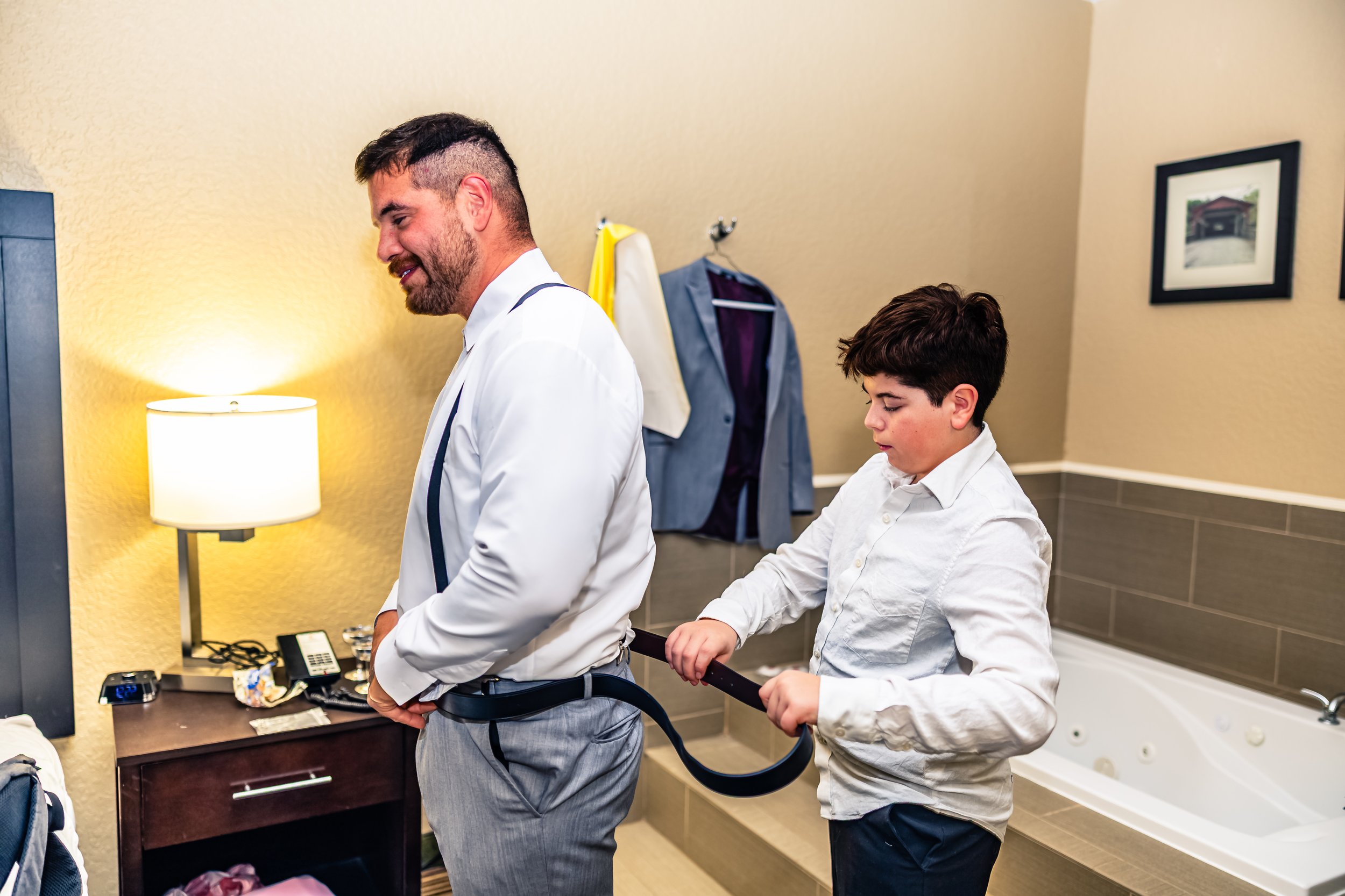 A man in a white shirt and gray pants is being helped to put on suspenders by a young boy in a white shirt, in a hotel room with beige walls, a lamp, a small dresser, and framed photos on the wall.