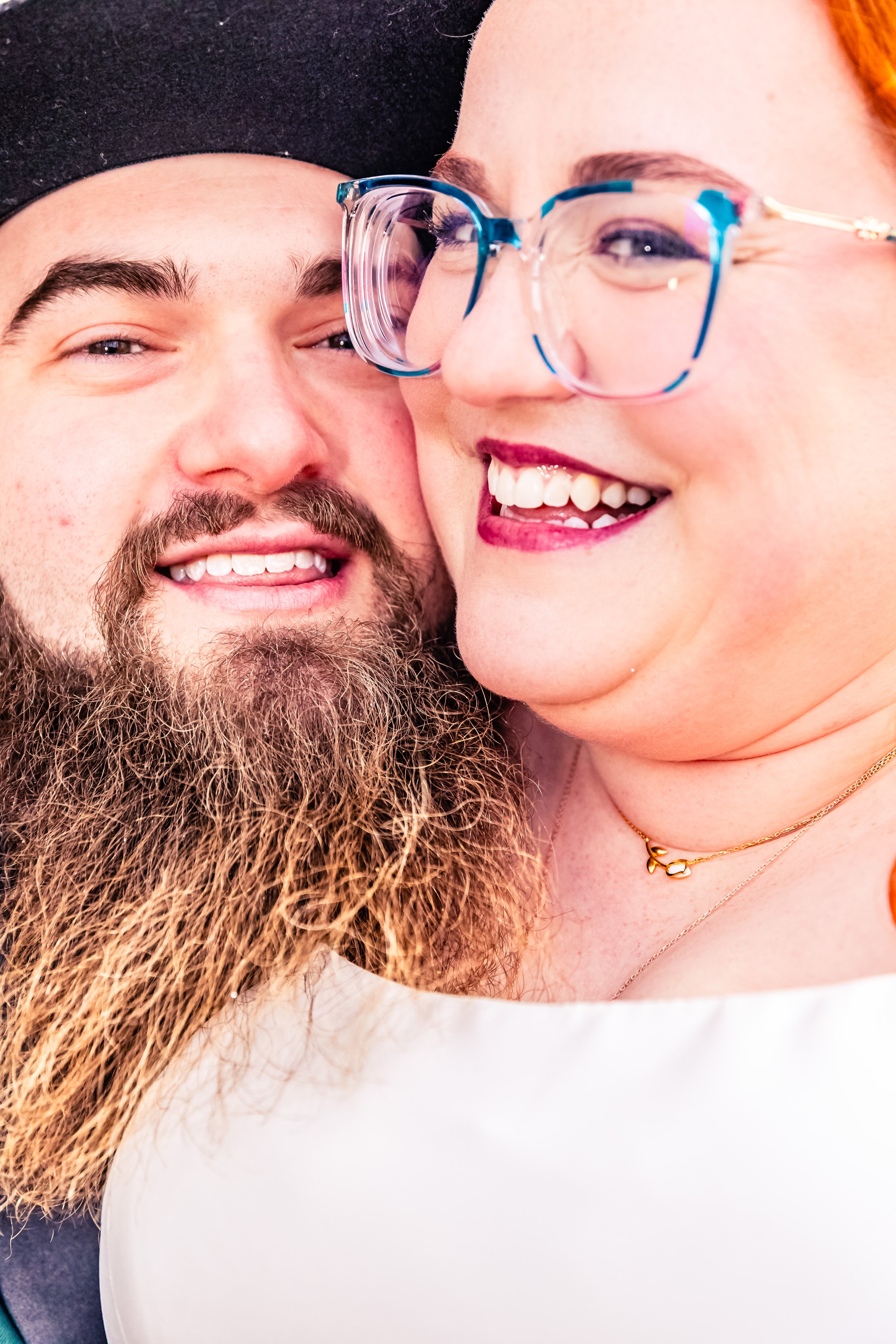 Close-up of a smiling couple, with the woman wearing glasses and jewelry, and the man with a beard and mustache, both showing happiness.