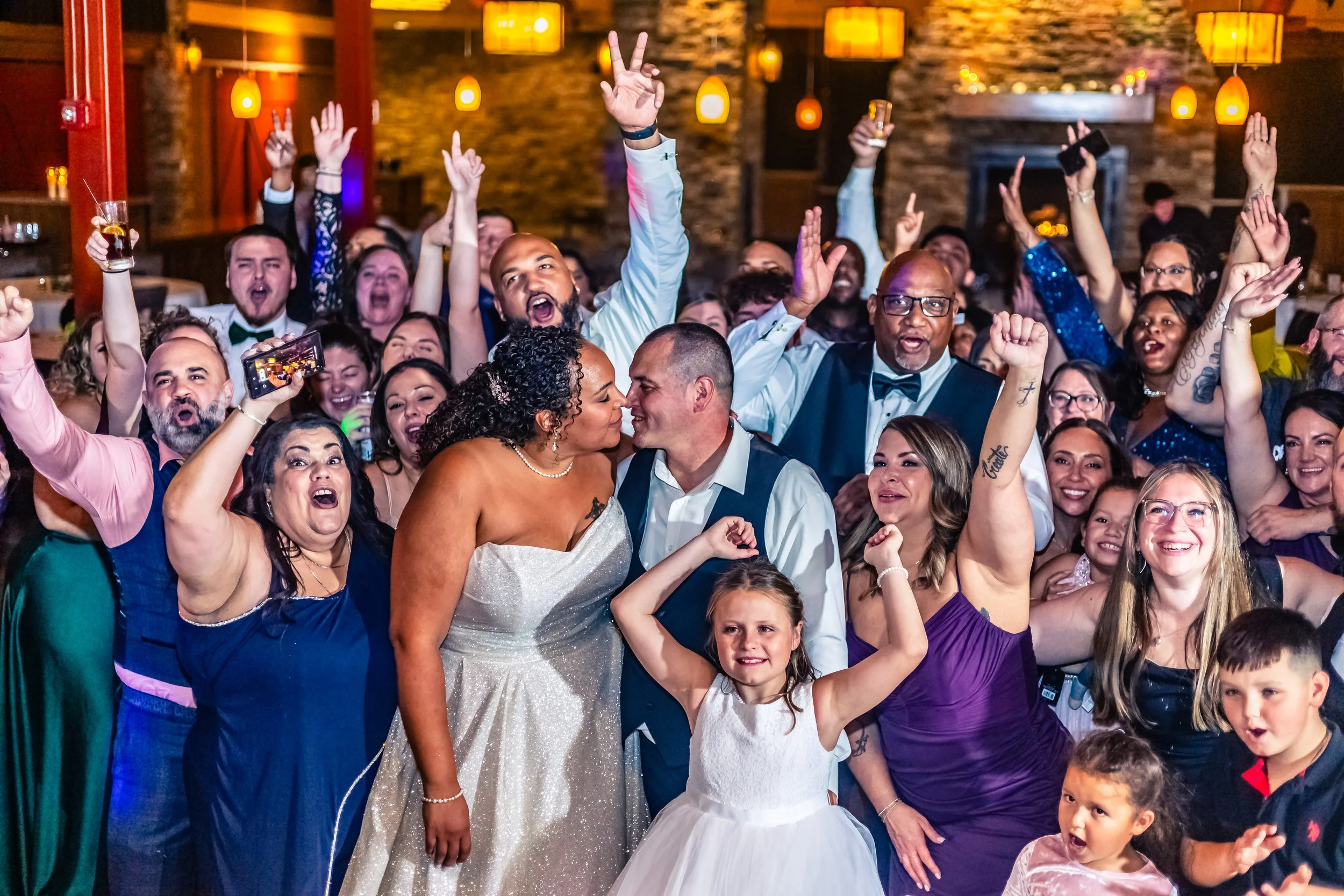 A wedding celebration with the bride and groom in the center surrounded by friends and family dancing and cheering.