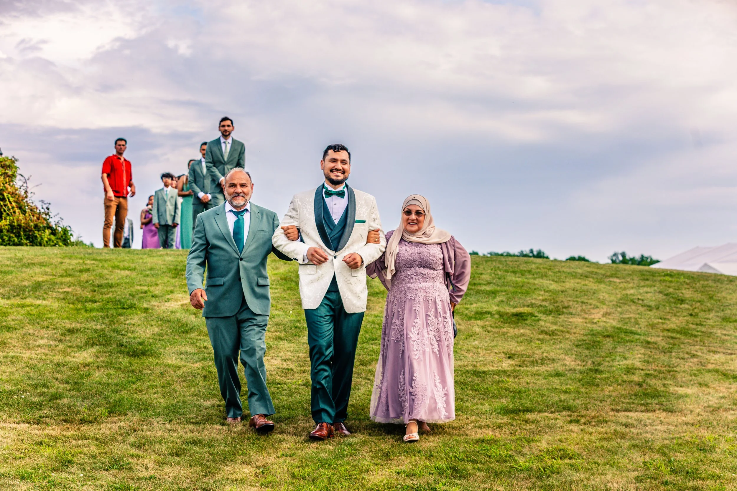 Group of people walking on a grassy hill, including a man in a white tuxedo with a teal bow tie, a woman in a lavender dress and hijab, and an older man in a teal suit, with more people in formal attire standing in the background under a partly cloud