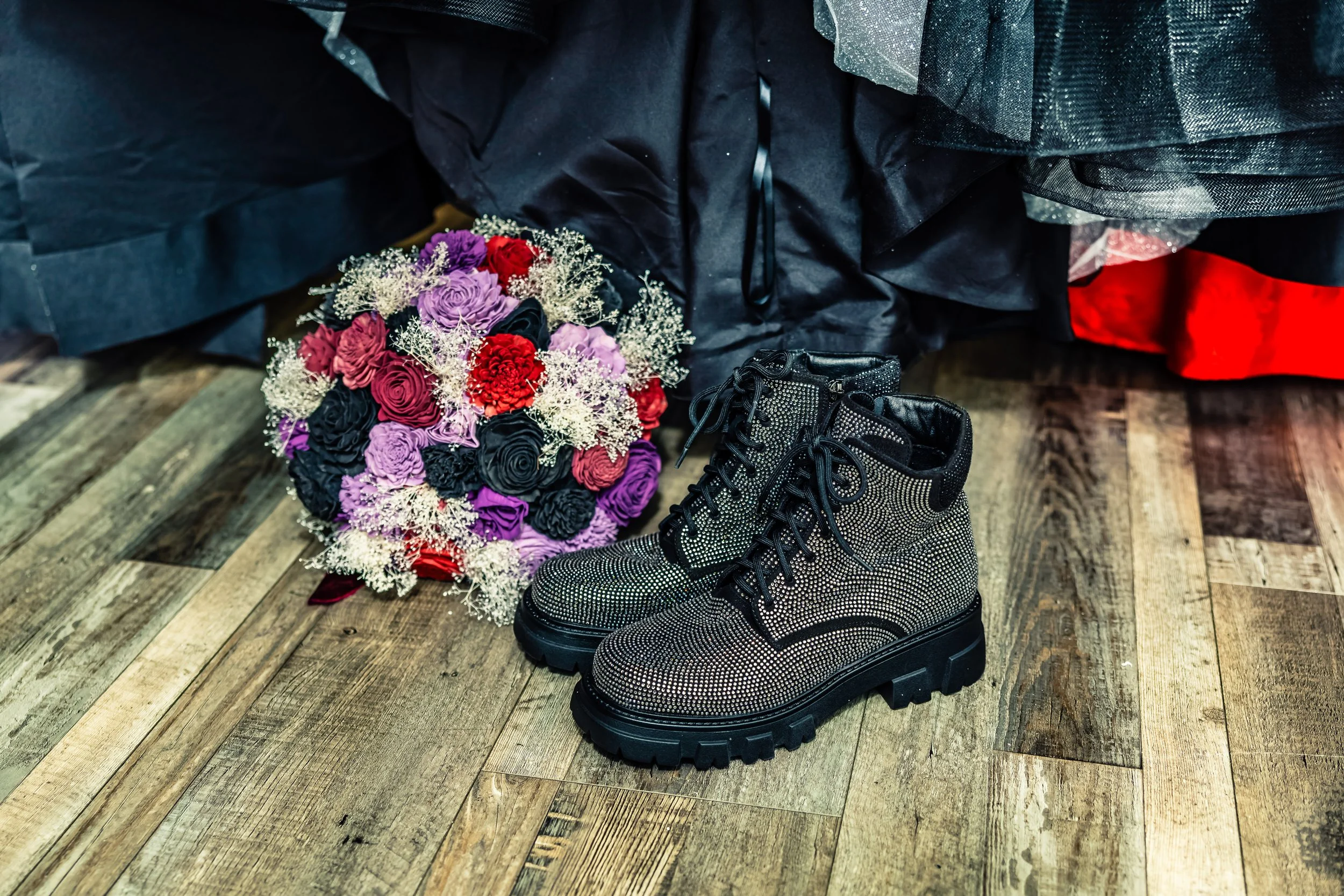 A bouquet of purple, red, and black roses with white baby's breath flowers, and a pair of black combat boots with rhinestones placed on a wooden floor near black and red fabric.