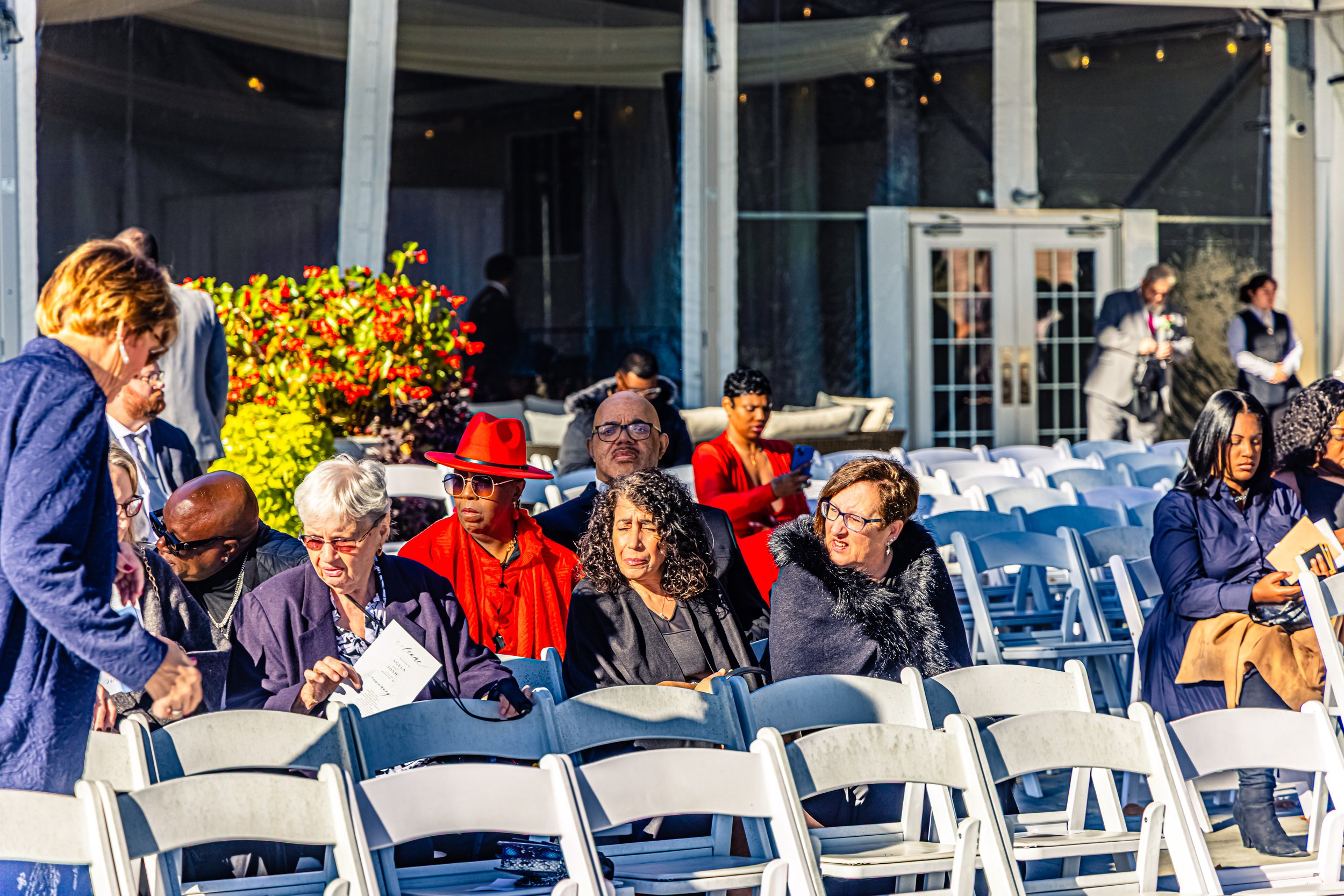 People sitting and standing in an outdoor event space with white chairs, some holding programs or devices, dressed in formal attire, with vibrant flowers and bright sunlight.
