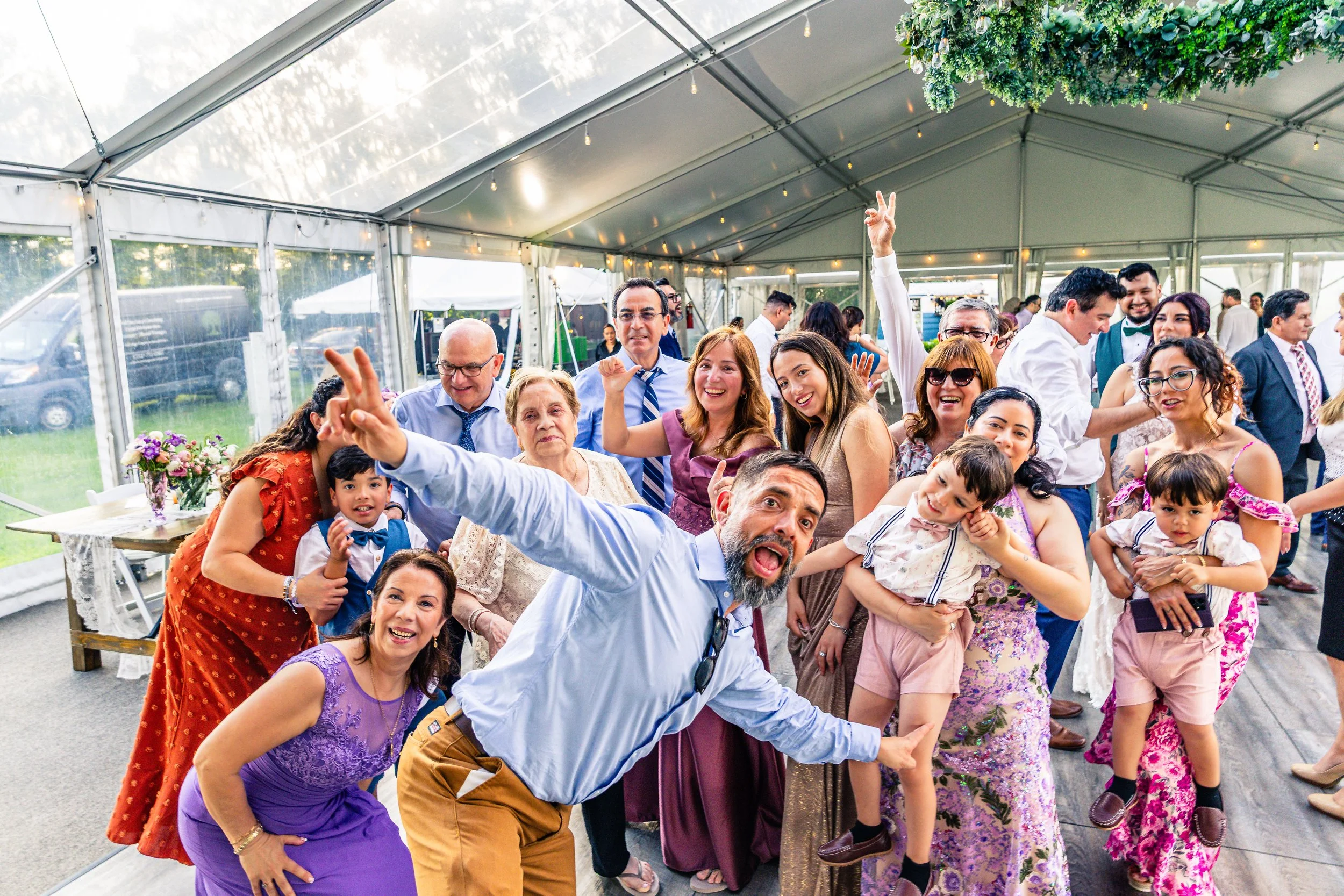 People dancing and celebrating at a wedding reception inside a large tent. There are both adults and children, with some making lively gestures and smiling.