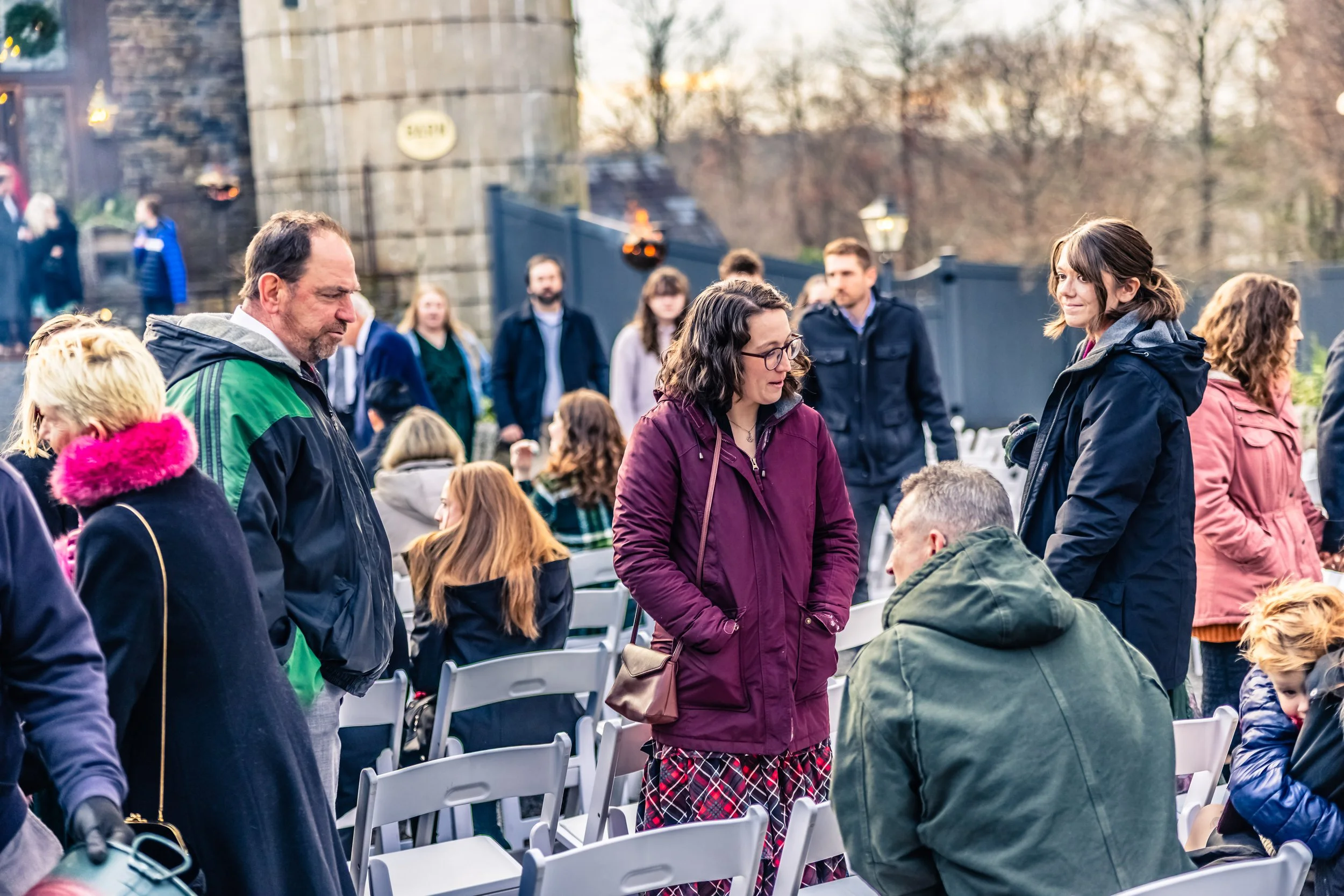 People gathered outdoors, some talking, sitting on chairs, while others stand, in a setting with stone structure and trees in the background during daytime.