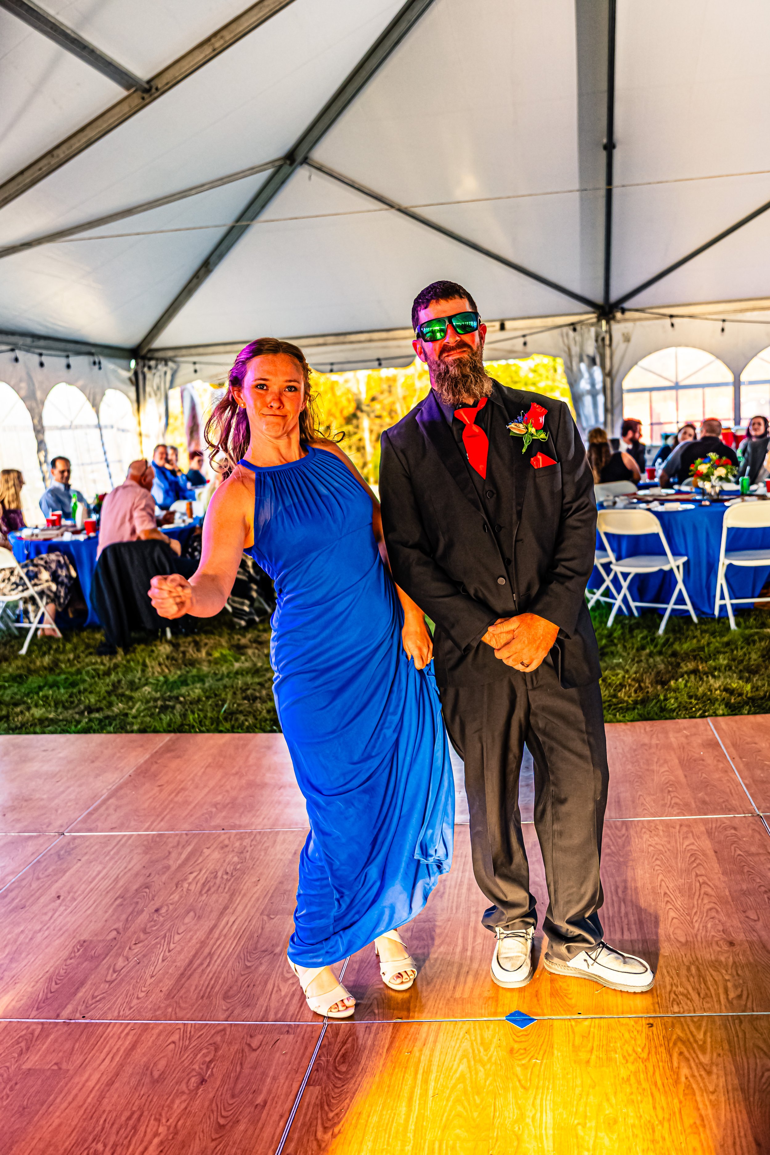 People dancing at a wedding reception under a large tent with blue tables and seated guests in the background.