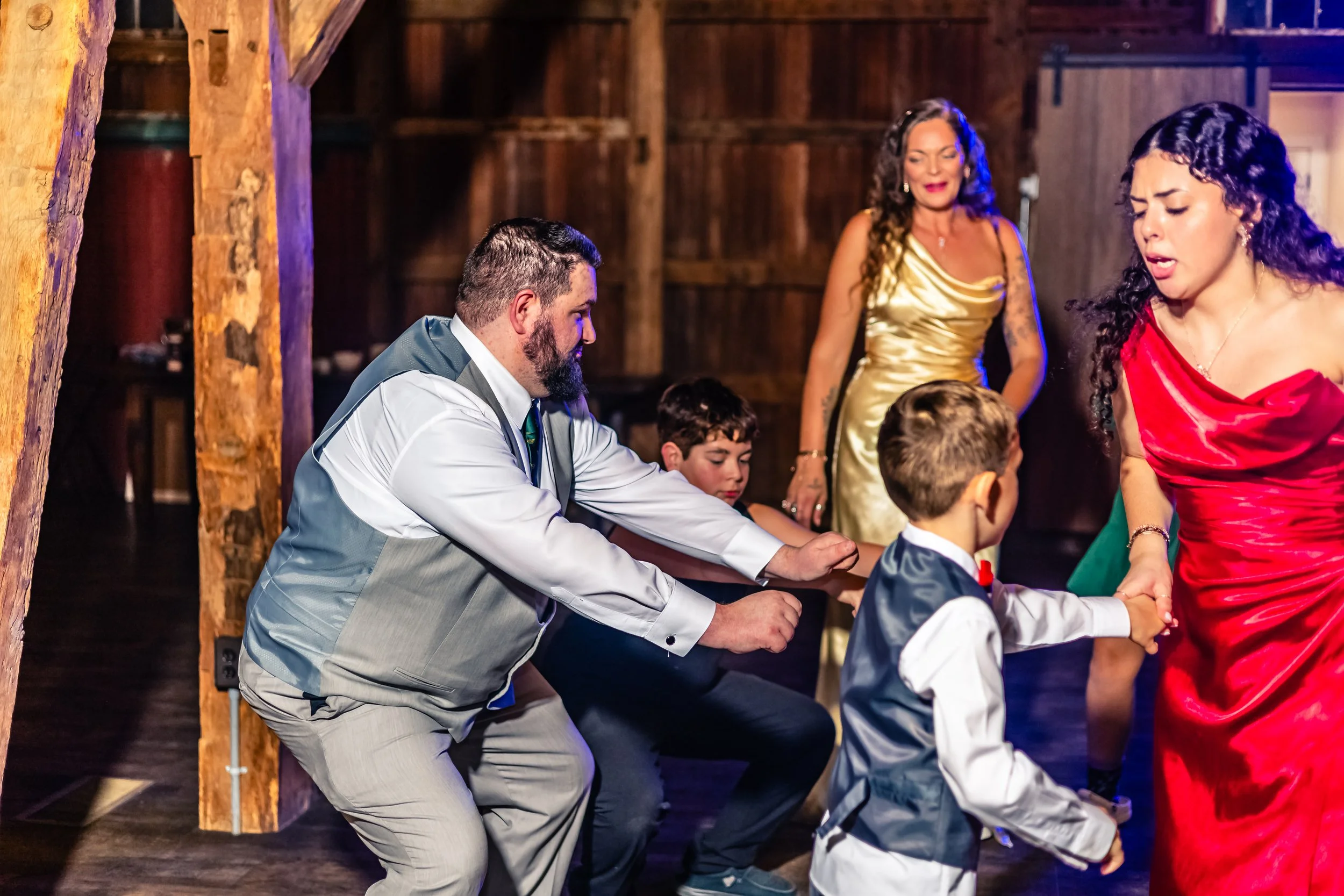 People dancing at a celebration, with a man in a light gray vest and white shirt holding hands with a woman in a red dress, and two children nearby, inside a wooden venue.