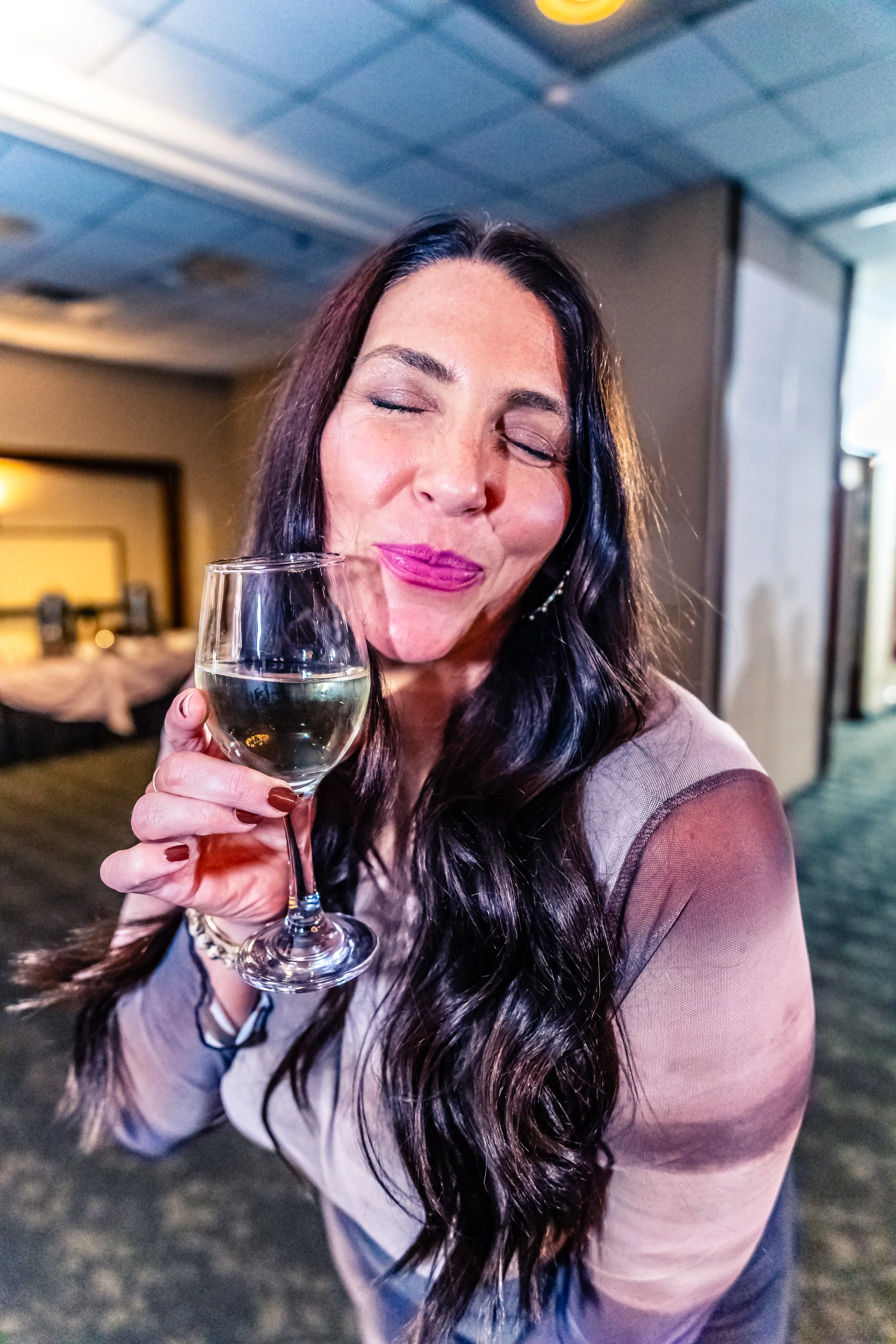 Woman with long dark hair, holding a glass of white wine, with her eyes closed and smiling in an indoor setting.