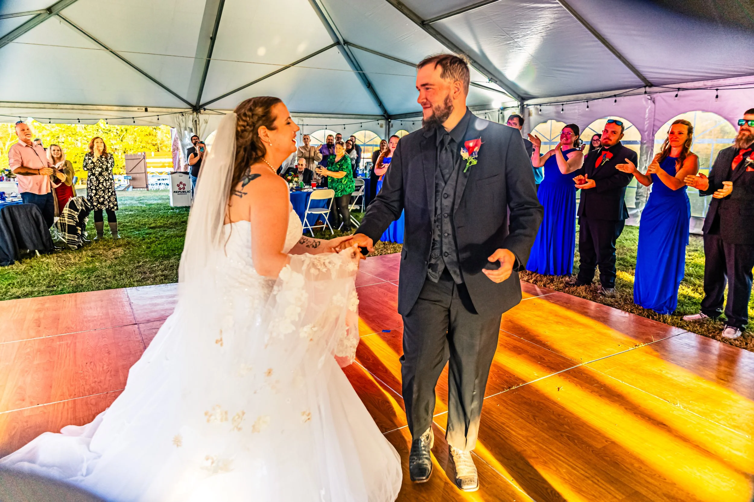 A bride and groom dance during their wedding reception inside a large white tent, surrounded by friends and family. The bride is wearing a white wedding gown, and the groom is in a dark suit. Guests are clapping and watching them dance.