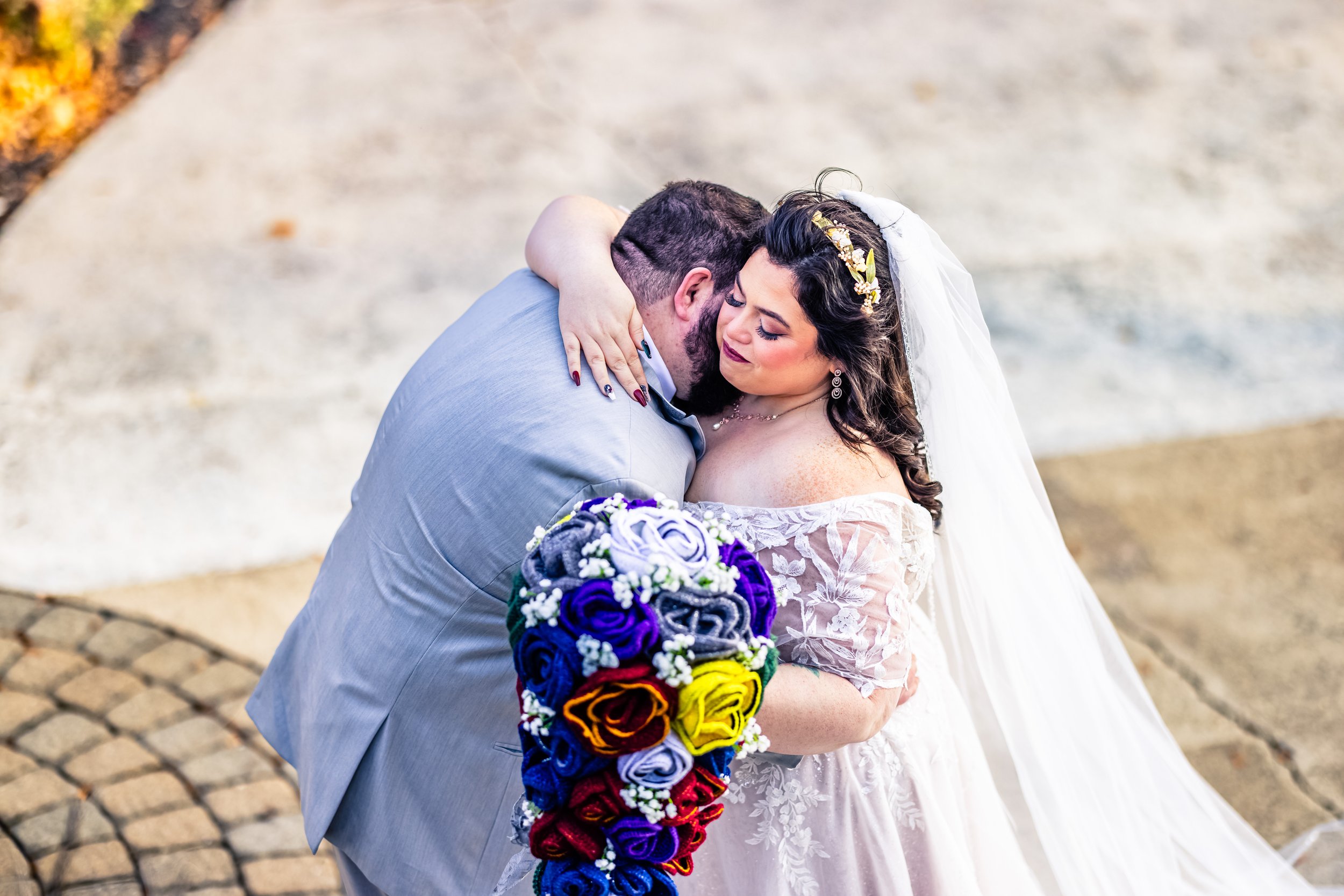 A bride and groom sharing a hug outdoors, with the bride holding a colorful bouquet of roses and wearing an off-the-shoulder lace wedding dress with a white veil.