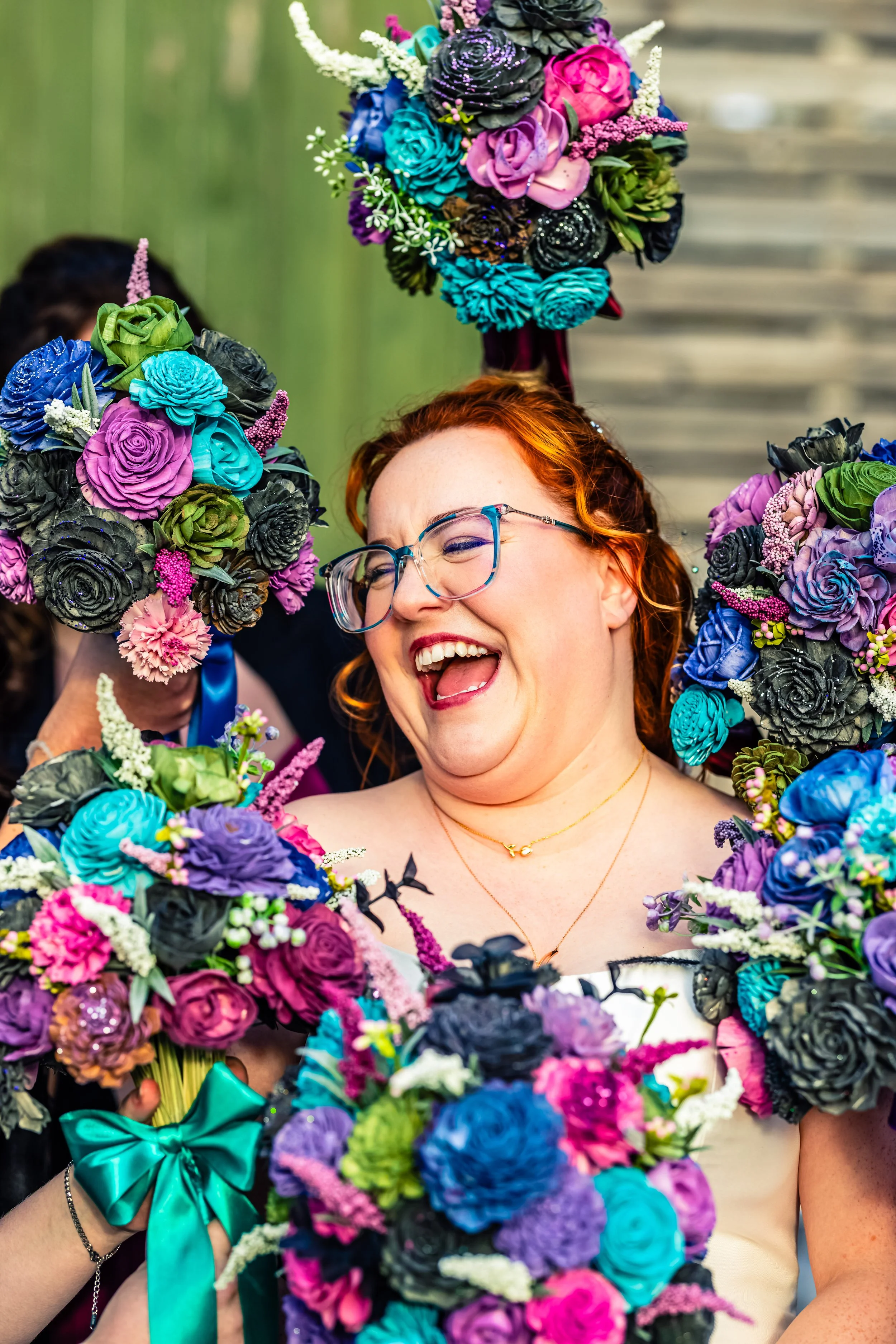 A joyful woman with glasses and red hair surrounded by colorful flower bouquets, smiling and expressing happiness.