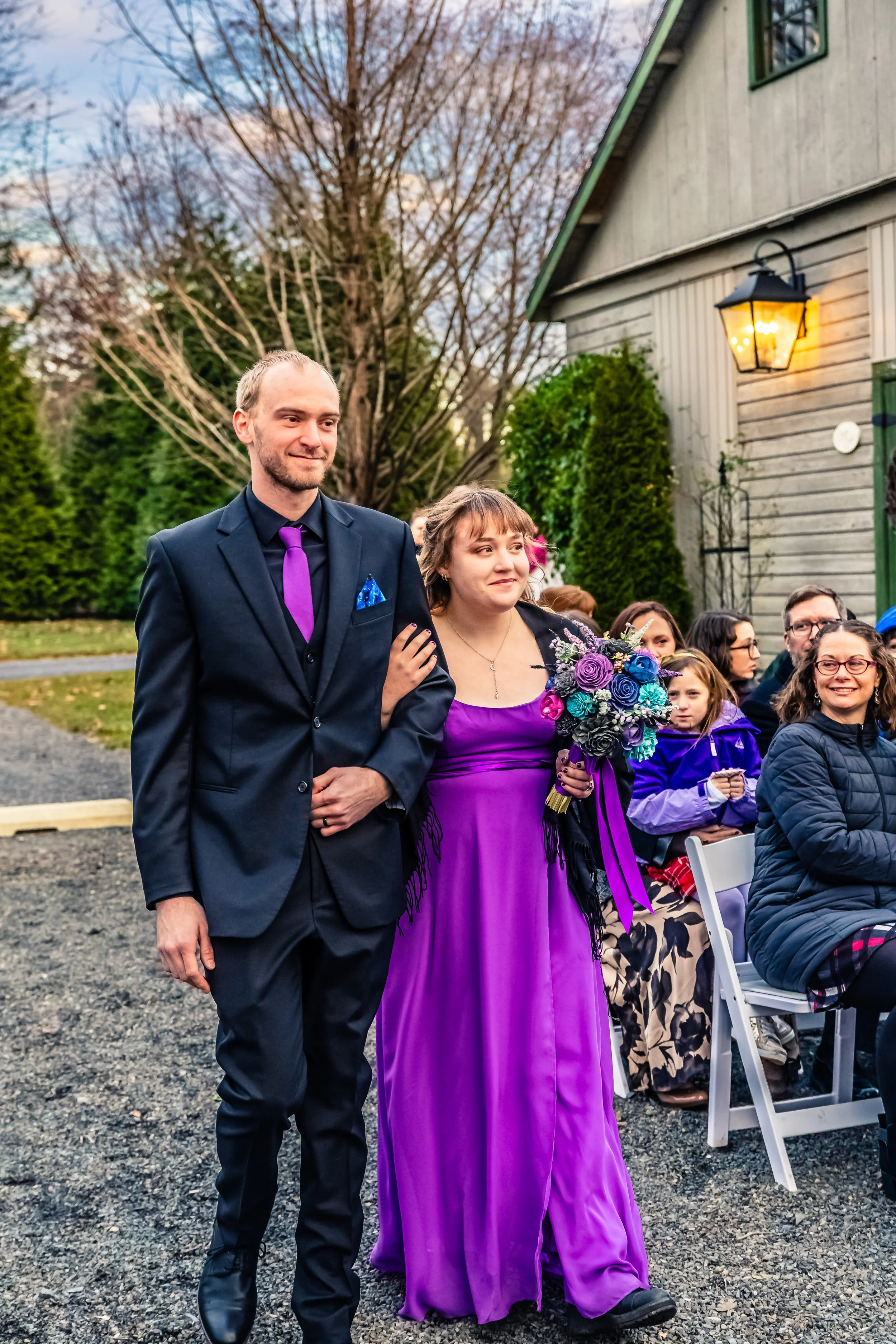 A woman in a purple dress holding a bouquet and walking arm in arm with a man in a black suit with purple tie at an outdoor wedding ceremony.