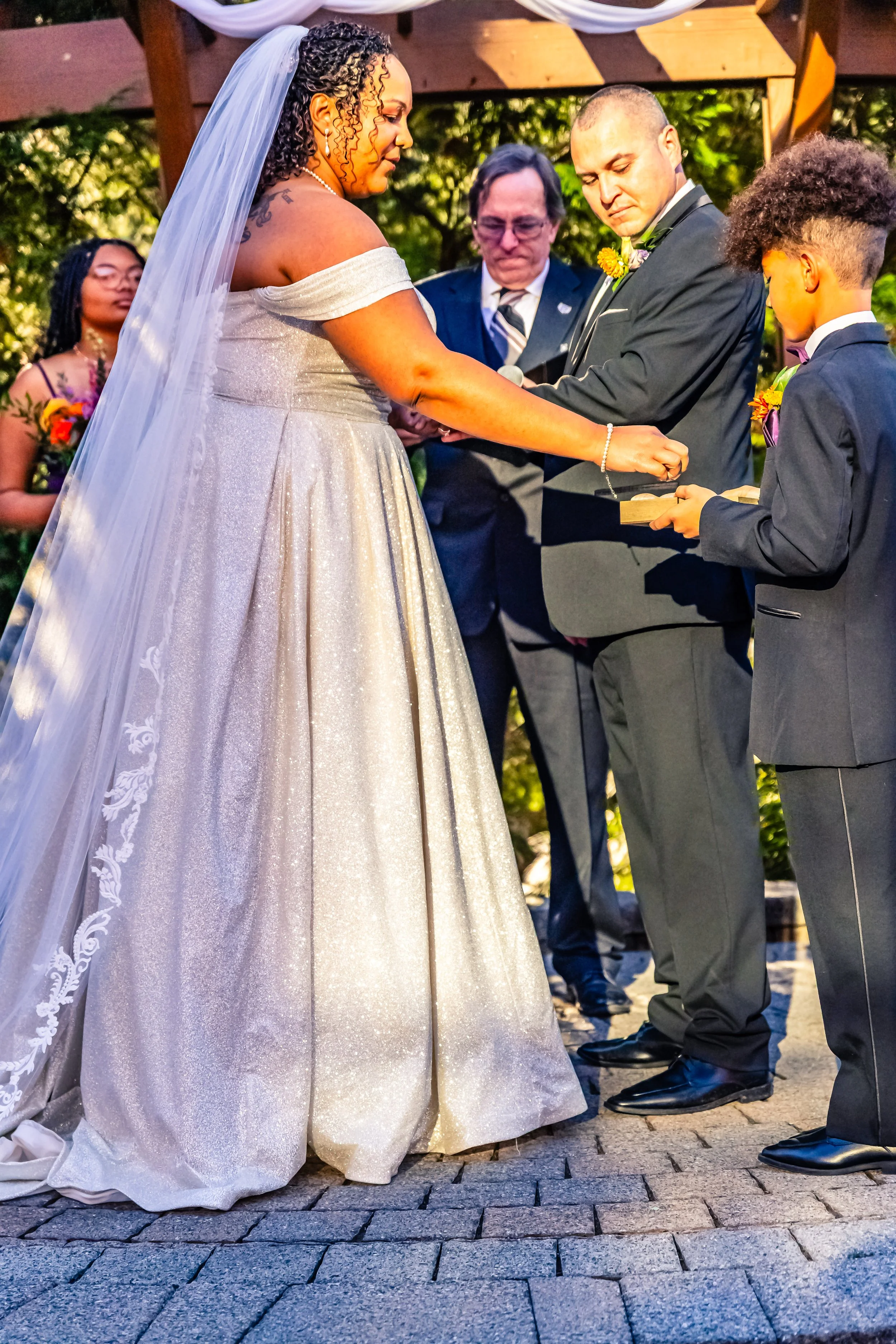 A bride and groom exchange rings during their outdoor wedding ceremony, with a officiant and two witnesses present.