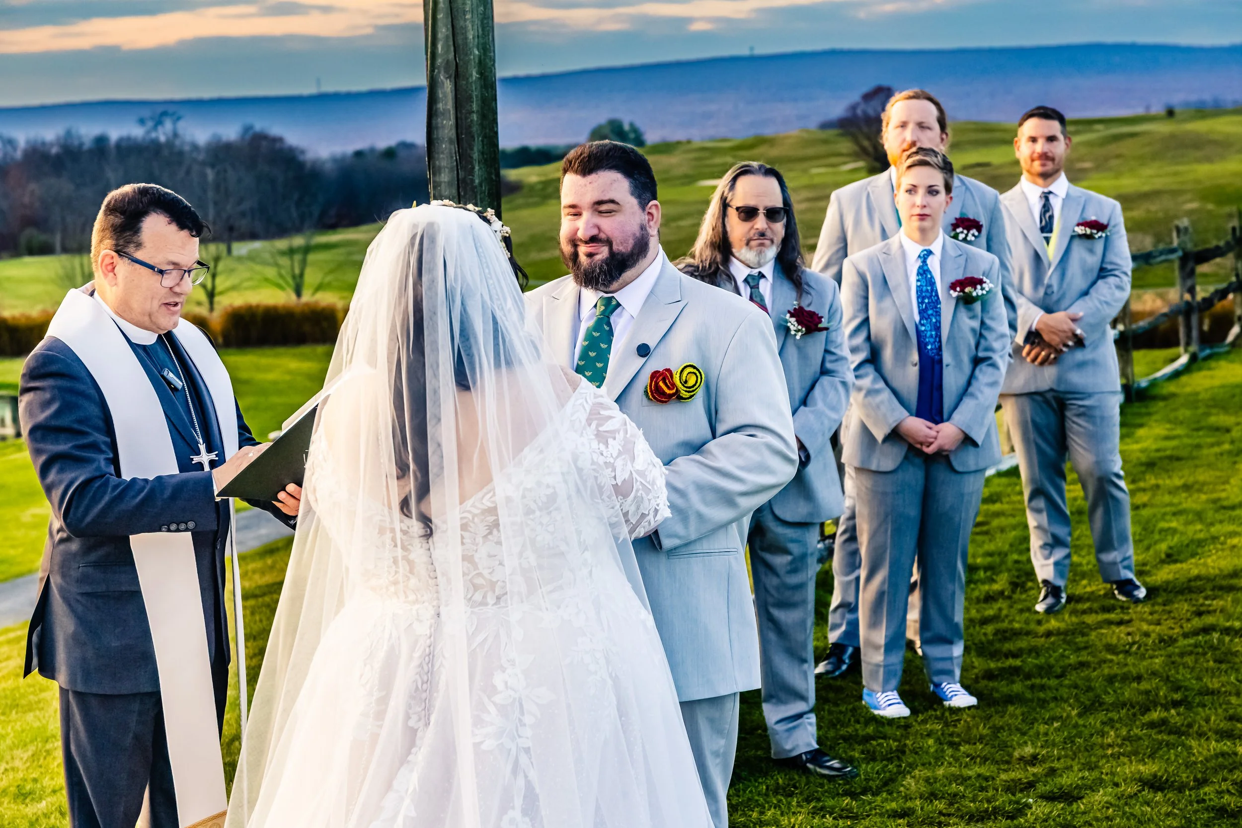 A wedding ceremony outdoors with a priest reading and a bride and groom exchanging vows, surrounded by five groomsmen in gray suits.