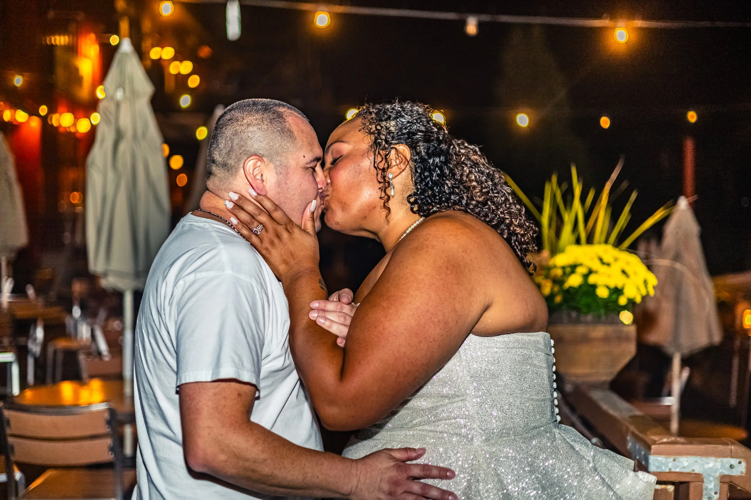 A woman and a man are kissing during a wedding reception at night, with string lights and umbrellas in the background.