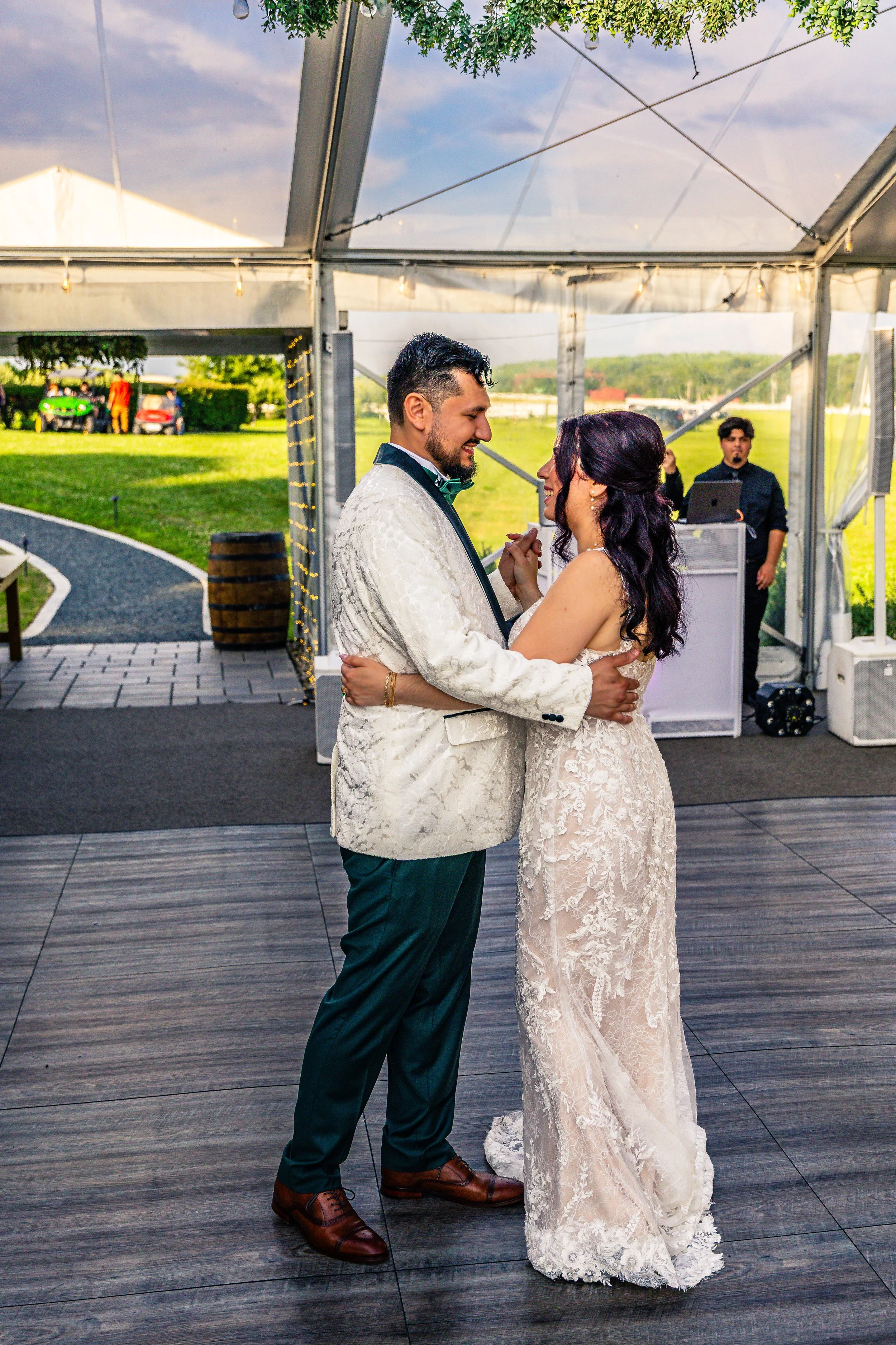 A newlywed couple sharing a dance under a clear tent, with a groom in a white tuxedo and a bride in a lace wedding gown, as a DJ plays music in the background during an outdoor wedding celebration.