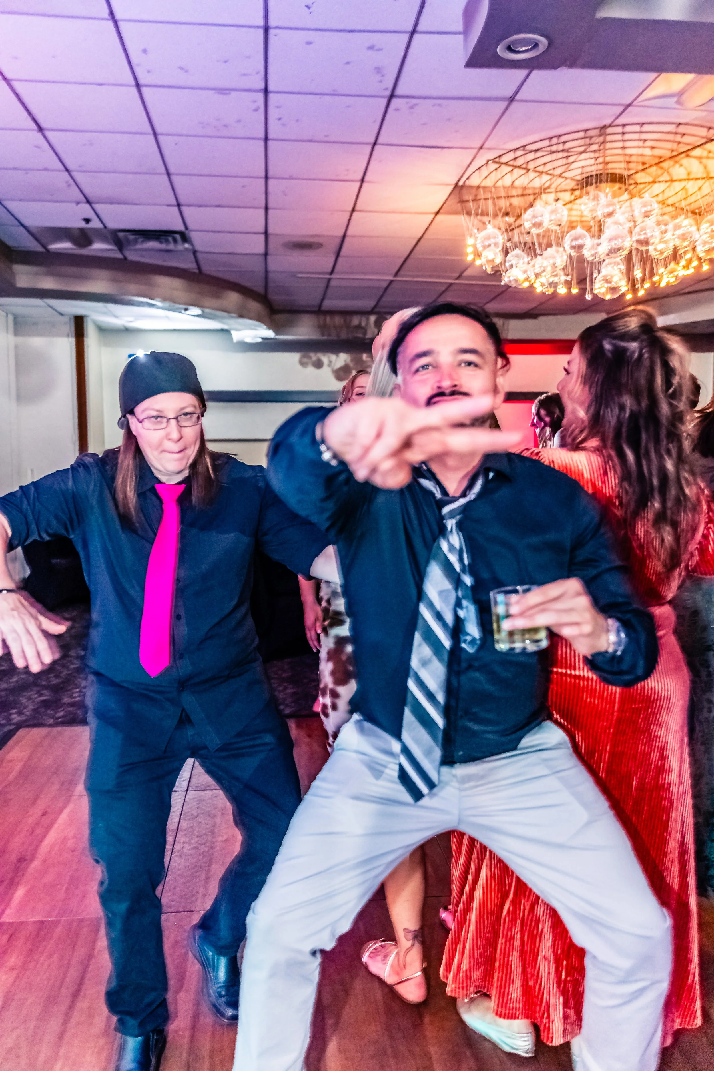 People dancing and having fun at a party, with one man in a black shirt and striped tie holding a drink in the foreground, and a woman in a red dress dancing behind him.