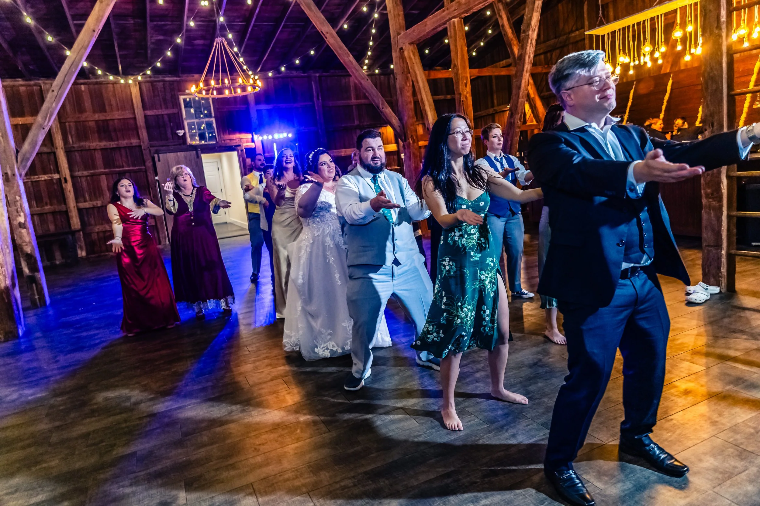 A group of people dancing and having fun in a rustic wooden barn decorated with string lights, during a wedding celebration.