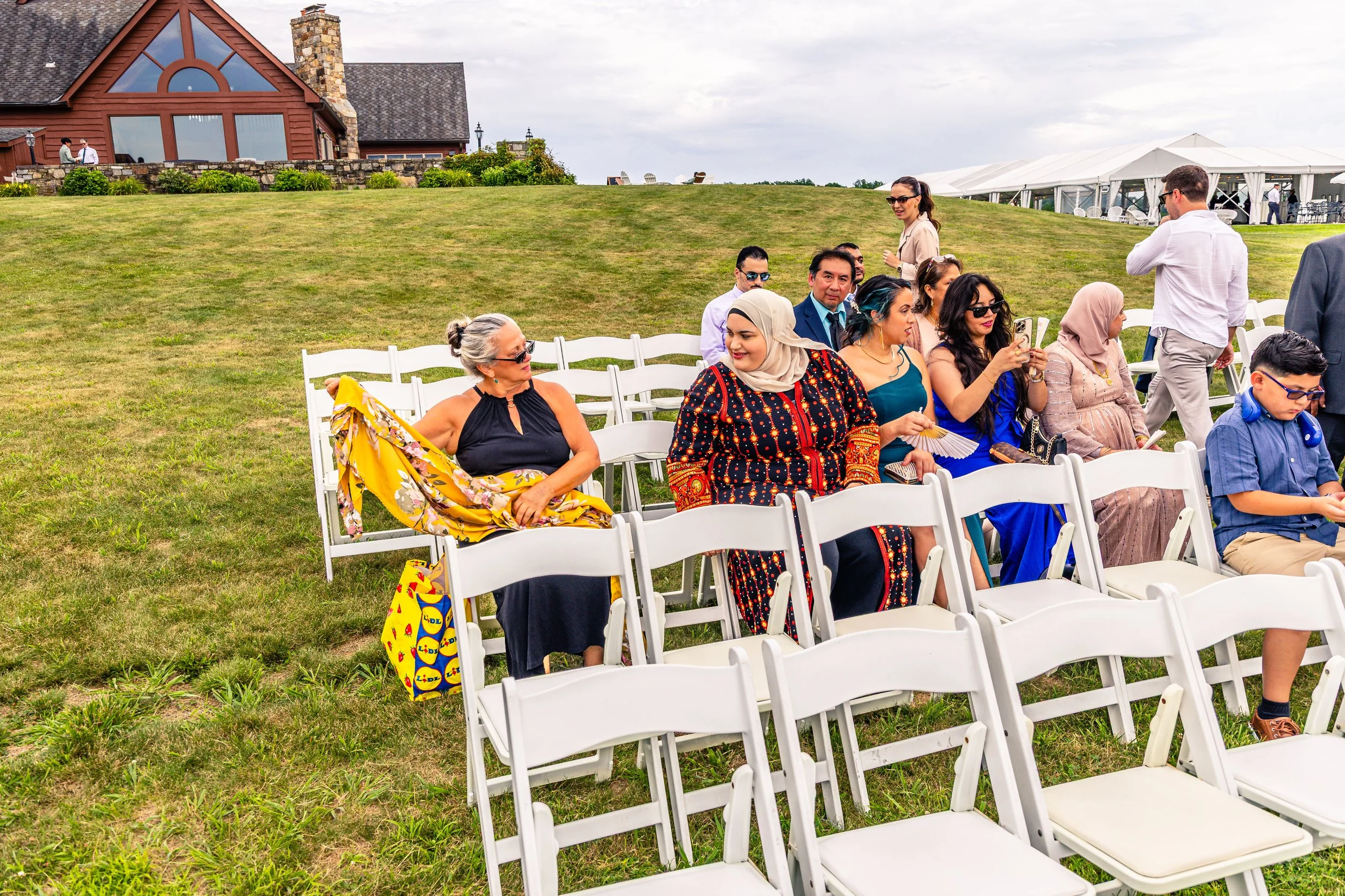People sitting outdoors on white folding chairs, attending an event on a grassy hill near a large house with a stone chimney, during daytime with cloudy sky.