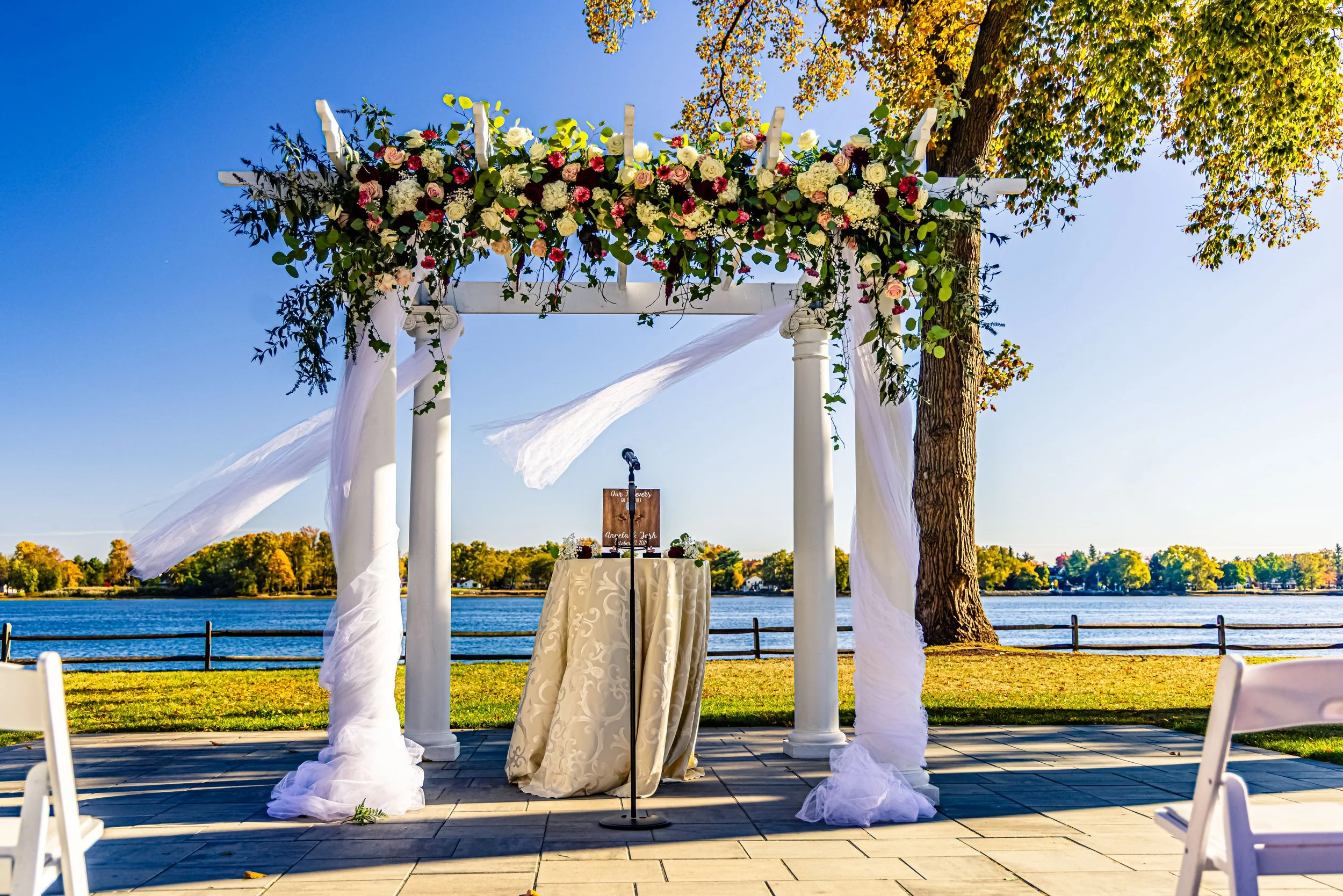 Wedding ceremony setup outdoors by a lake with a decorated archway of white columns and floral arrangements, white fabric draped around the columns, with a small table and microphone in front, trees with fall foliage in the background.