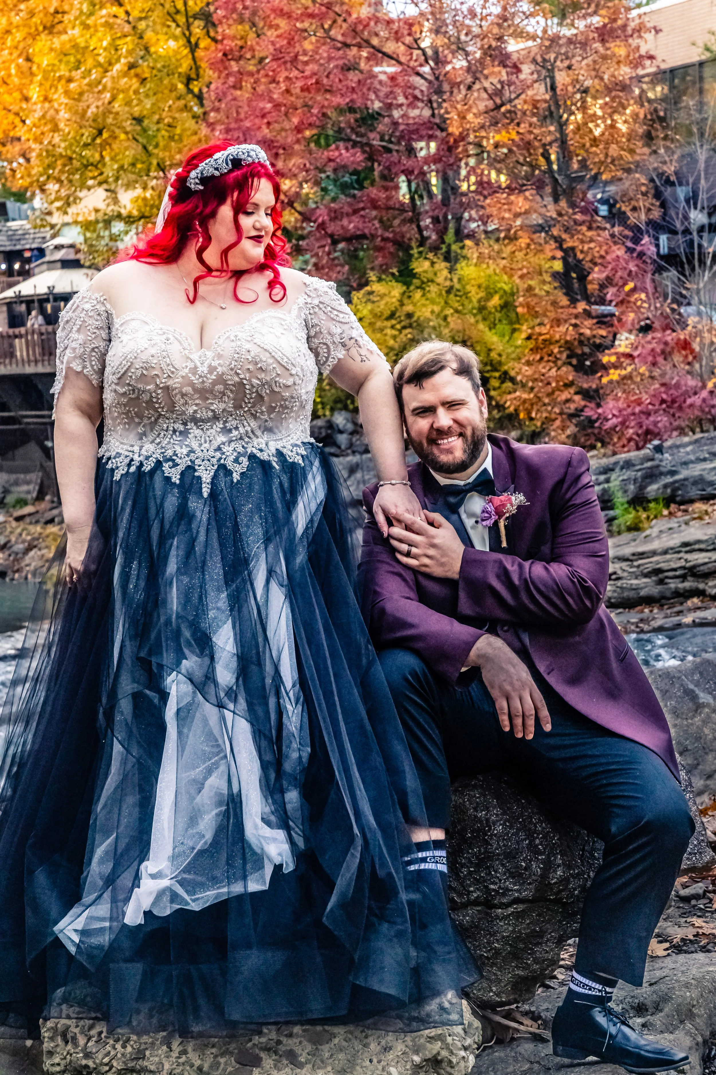 A woman and a man dressed in formal wedding attire outdoors near a river with autumn-colored trees in the background. The woman has red hair, a tiara, and a wedding gown with intricate lace and a dark blue tulle skirt. The man has a beard and is wear