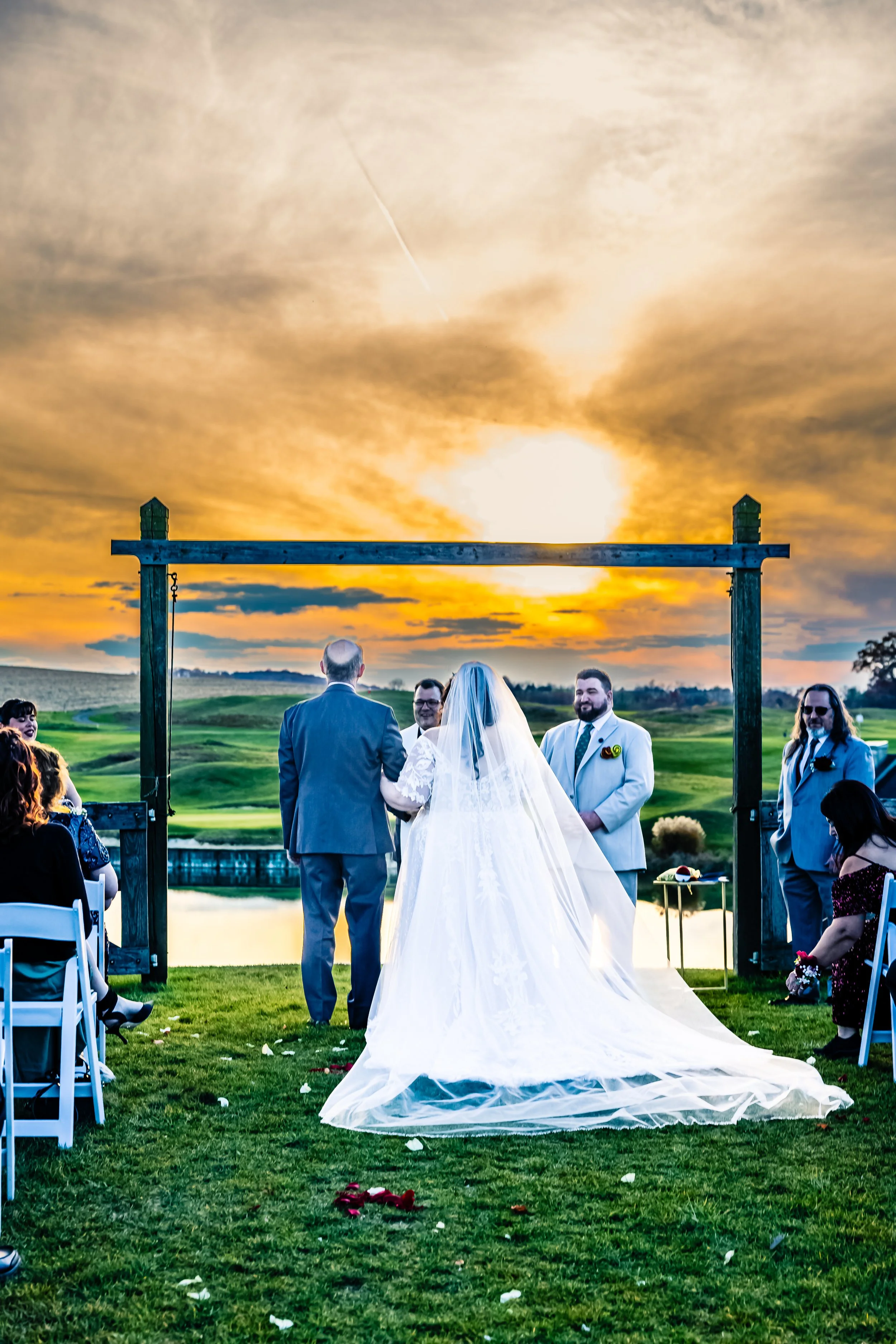 A wedding ceremony taking place outdoors at sunset, with a bride and groom standing under a wooden arch, surrounded by seated guests beside a lake and green landscape.