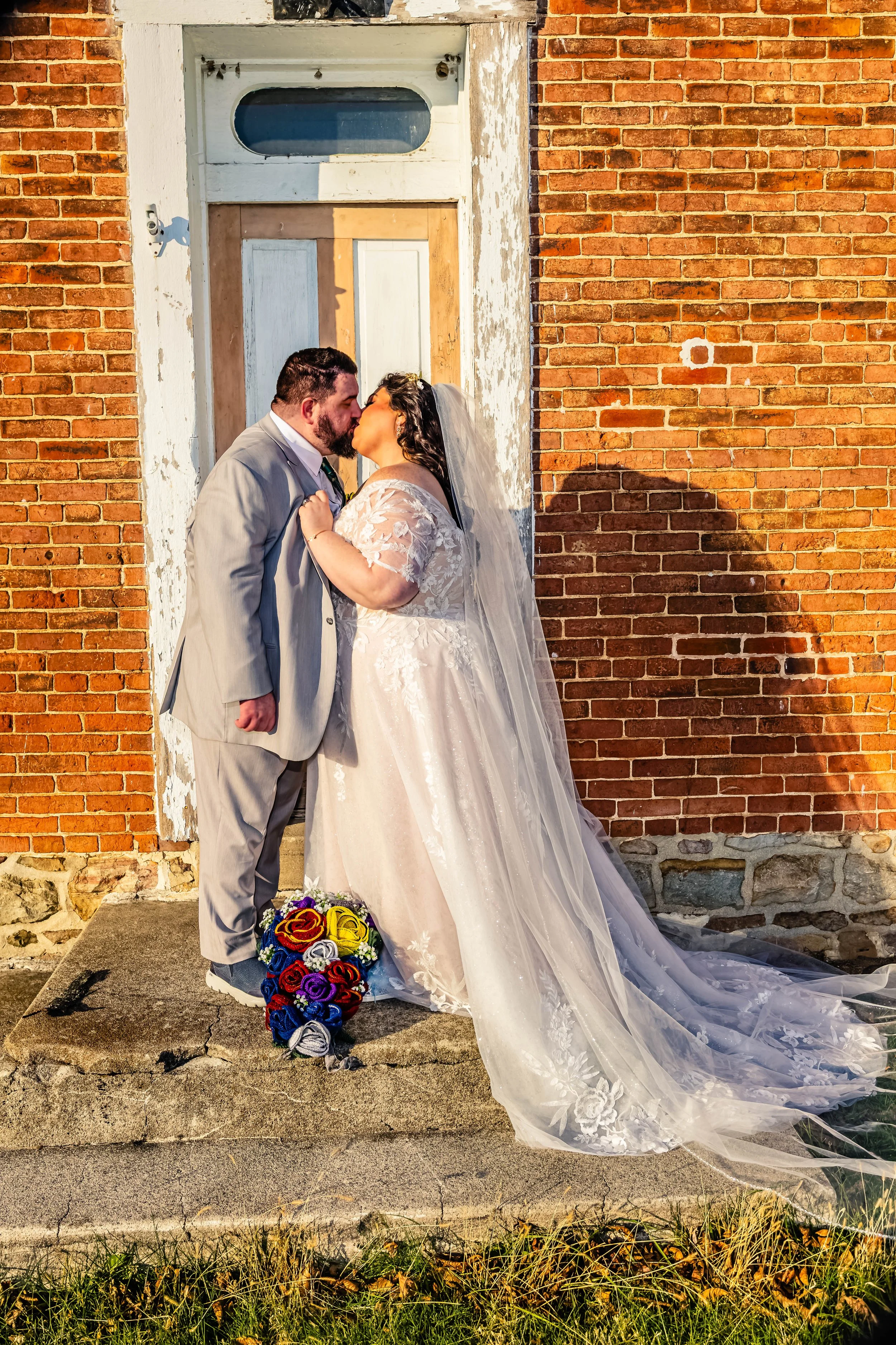 A bride and groom kiss outside a brick building, standing on concrete steps with a colorful bouquet on the ground nearby.