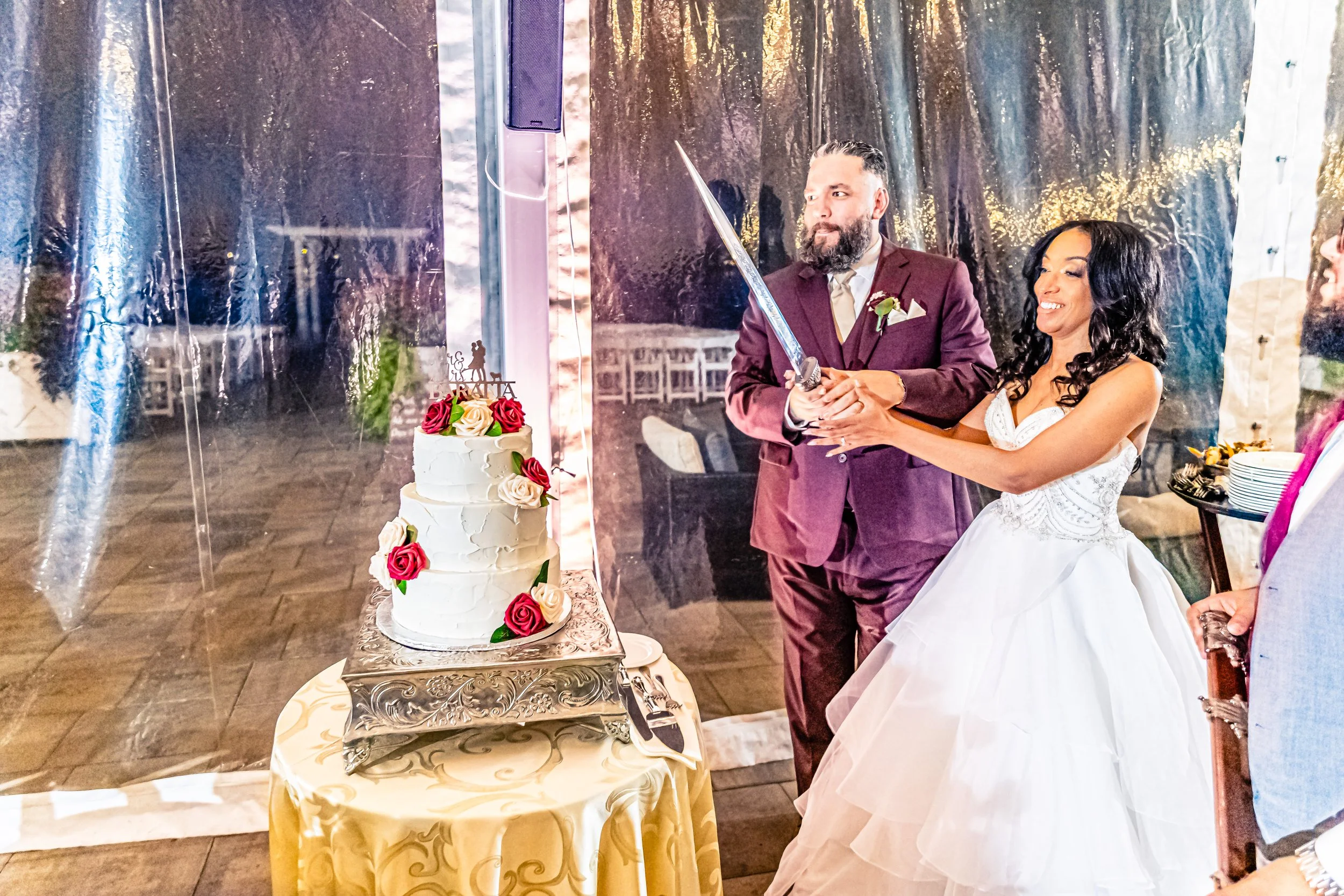 Bride and groom cutting a wedding cake with a sword, bride wearing a white wedding gown and groom in a purple suit, wedding cake decorated with roses, wedding reception setting.