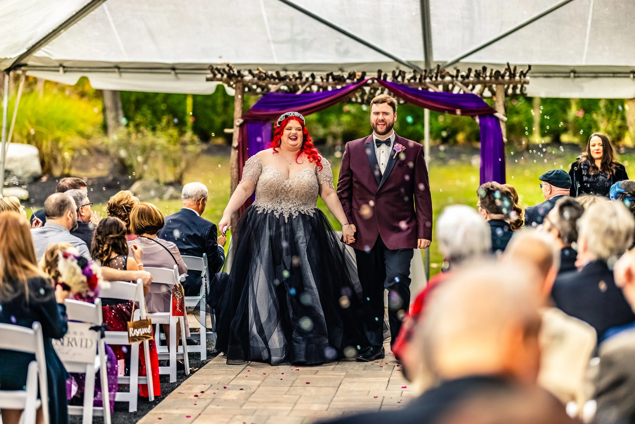 A bride with red hair in a beige and black wedding gown and a groom in a maroon tuxedo walk hand-in-hand down the aisle of an outdoor wedding ceremony under a canopy with purple drapery, surrounded by seated guests, some clapping and celebrating.