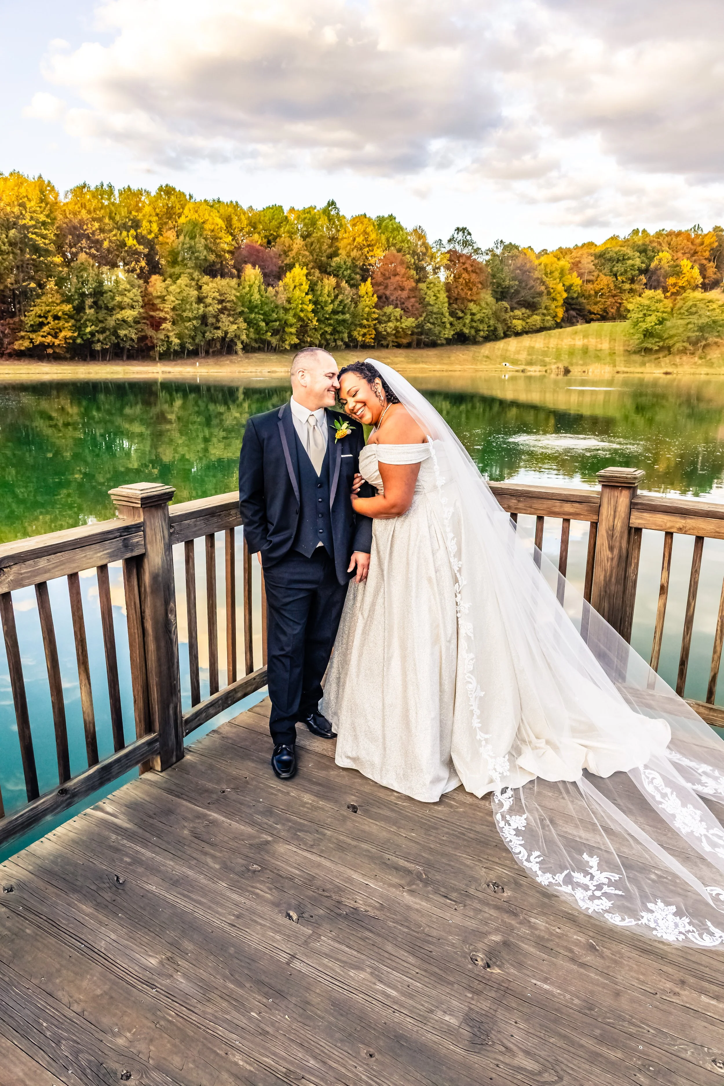 A bride and groom sharing a joyful moment on a wooden dock by a lake, with autumn trees and a cloudy sky in the background.