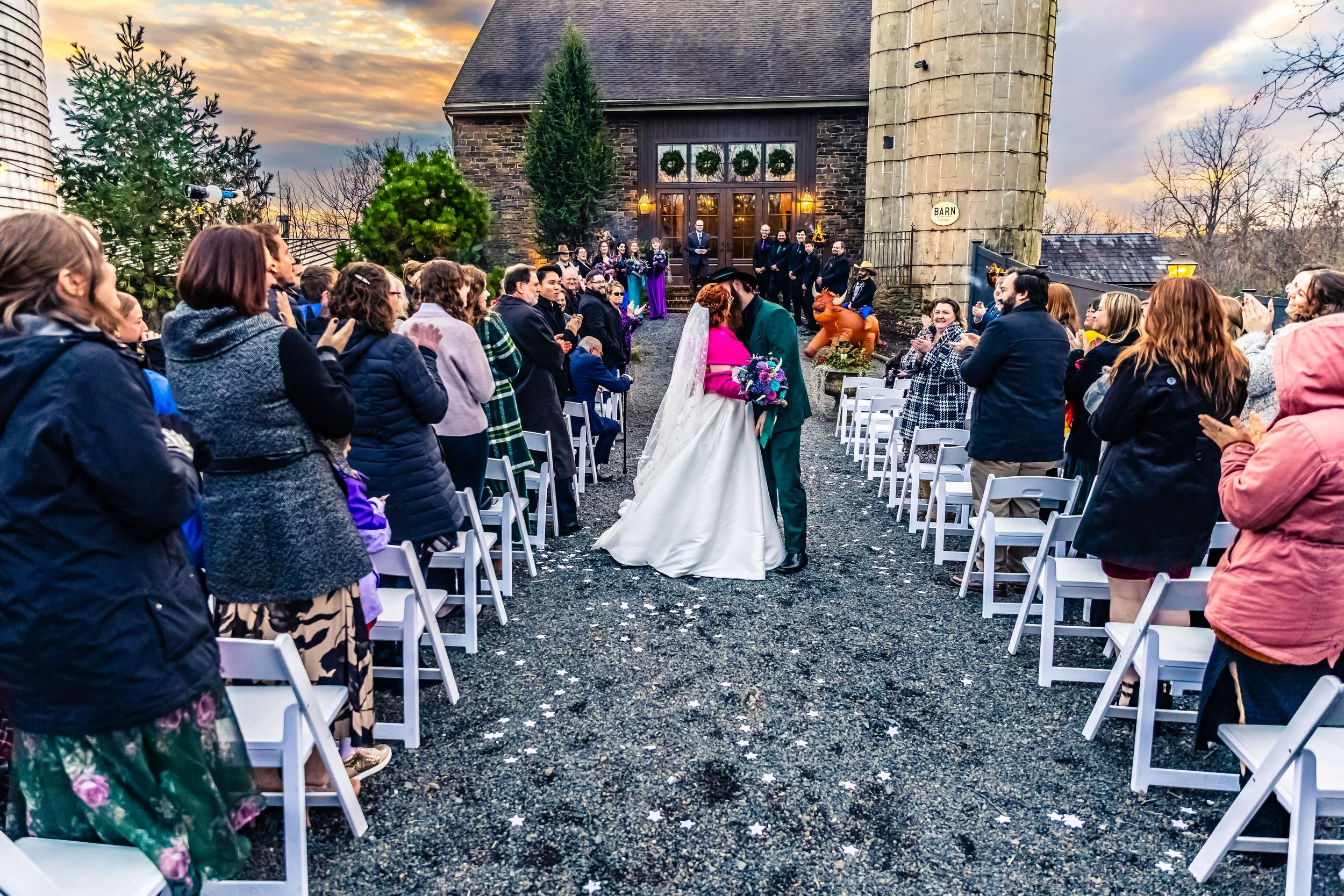 A wedding ceremony taking place outdoors in front of a rustic building during sunset. The bride and groom are sharing a kiss, surrounded by guests standing and applauding. The scene includes festive decorations and a cozy atmosphere.