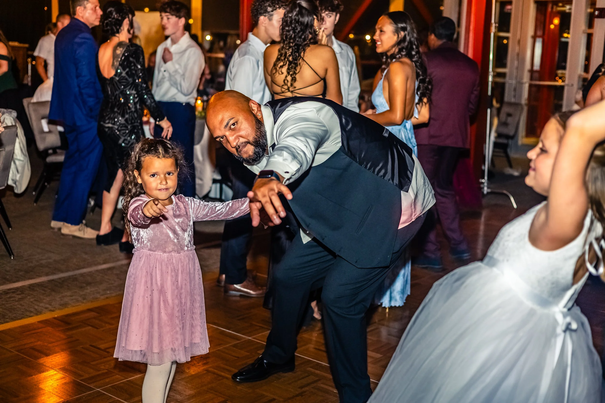 A man in a suit and a little girl in a pink dress dance happily together at a social gathering or party. Other people are in the background, some dancing and some chatting, with a lively atmosphere.