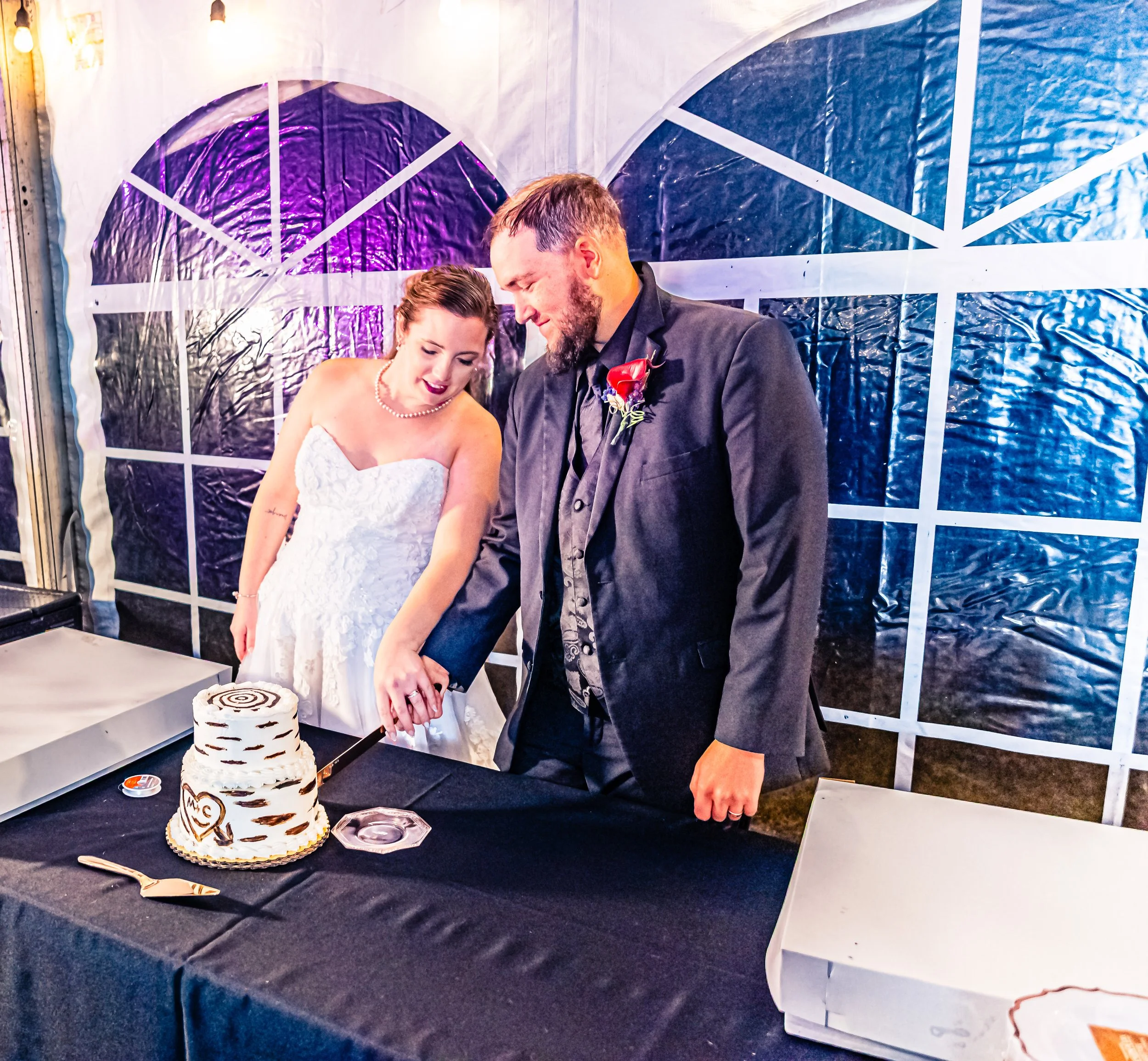 A bride and groom cutting their wedding cake together at a reception. The bride is in a strapless white wedding dress and the groom is in a dark suit with a red boutonniere. They are smiling and looking at the cake, which is decorated with black and 
