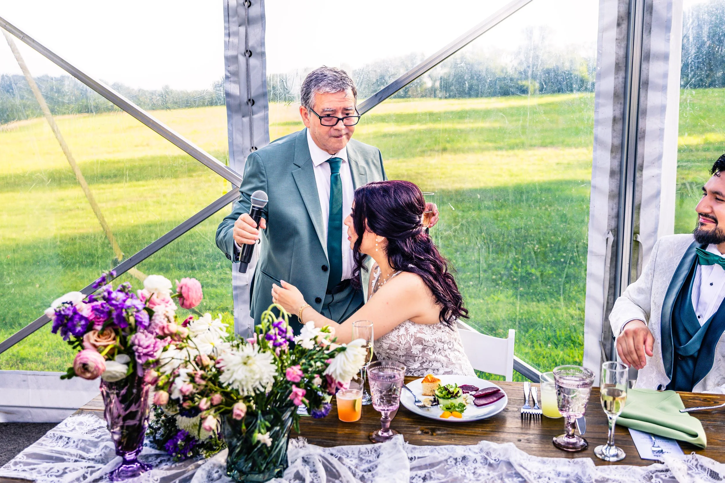 An elderly man in a teal suit holding a microphone, likely giving a speech at a wedding, while a woman in a wedding dress leans in to him with her hands on his chest. A man in a white tuxedo with a green bow tie is sitting at the table, smiling. The 