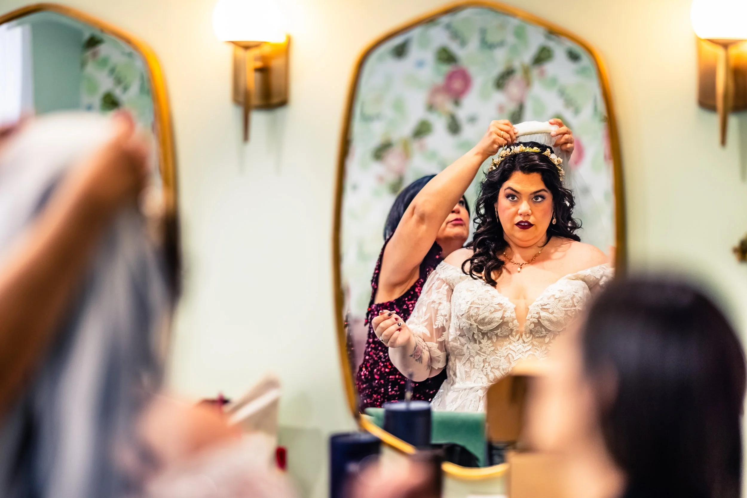 A woman in a wedding dress is having a floral crown placed on her head by another woman, seen through a mirror.
