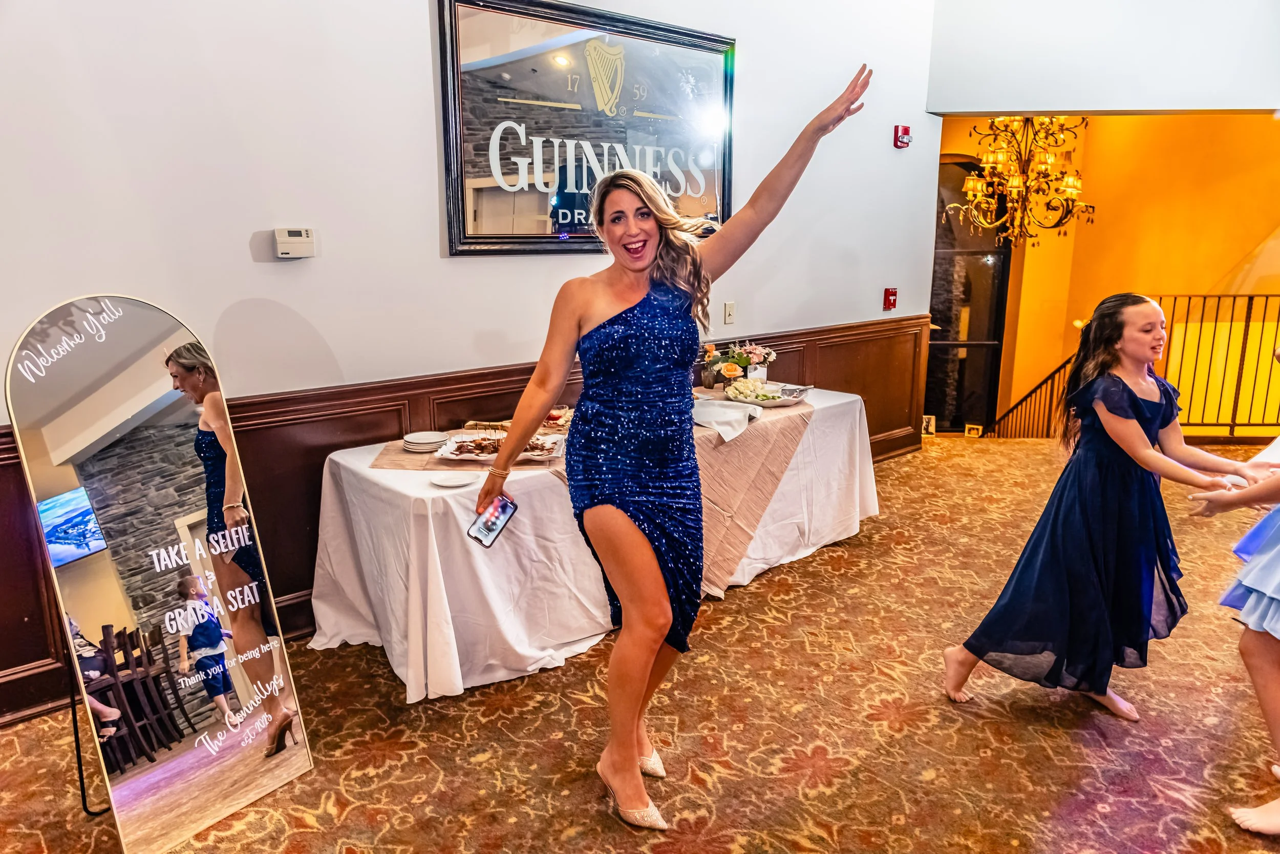 Woman in a blue sequined dress with a thigh-high slit, dancing and smiling in a decorated event space, with children in blue dresses, a buffet table, and a large mirror with instructions to take a selfie.