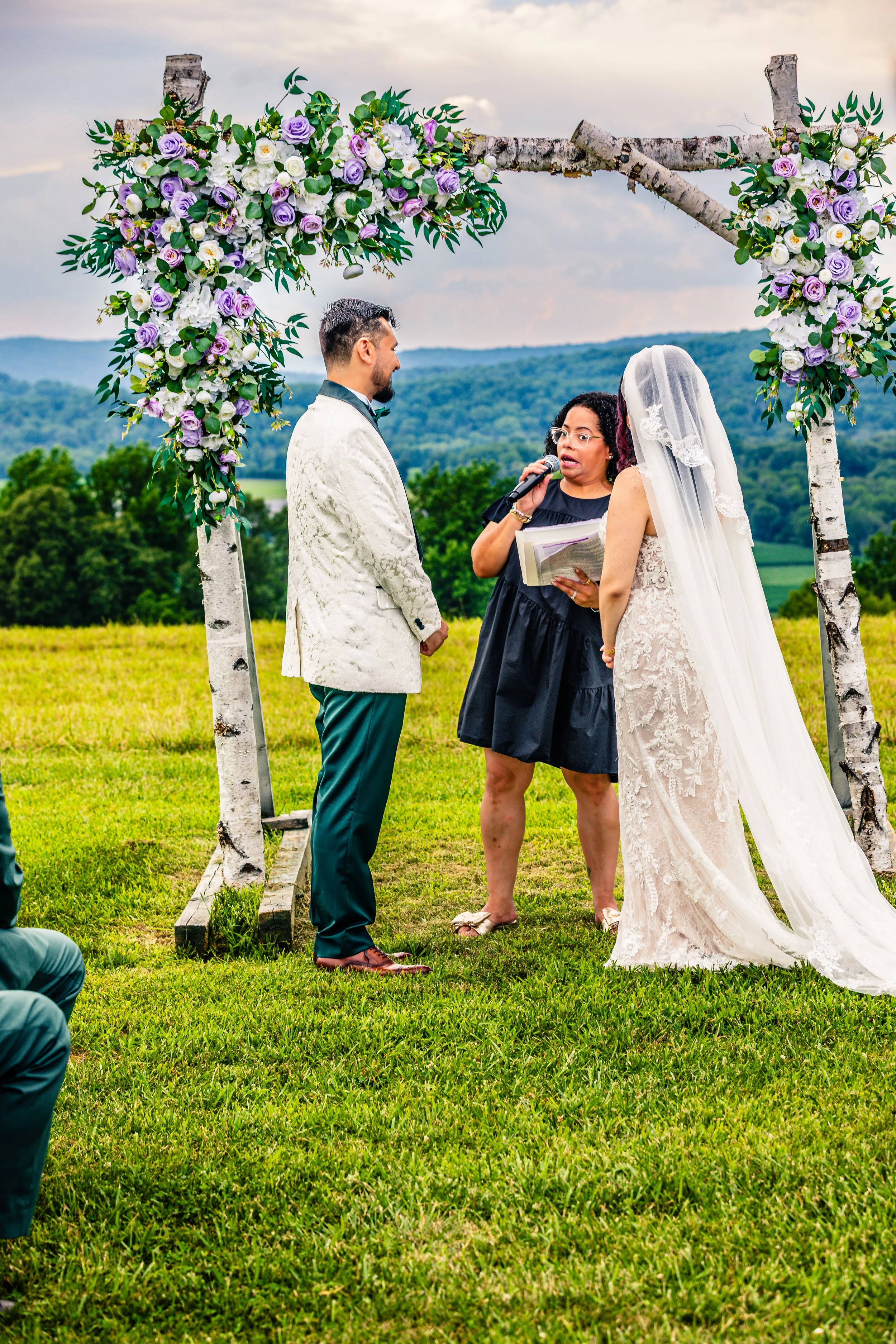 A couple getting married outdoors beneath a floral arch, with an officiant reading vows, on a grassy field with a backdrop of rolling hills and cloudy sky.
