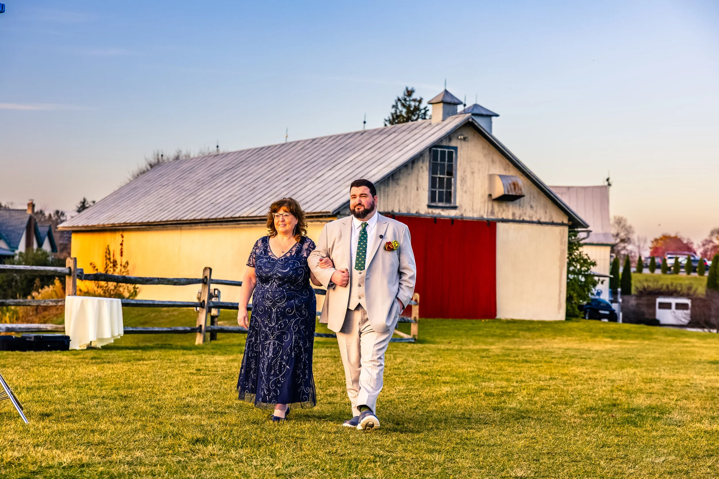 A man in a light beige suit and a woman in a navy blue dress walking together on a green lawn during sunset, with a white and red barn in the background.