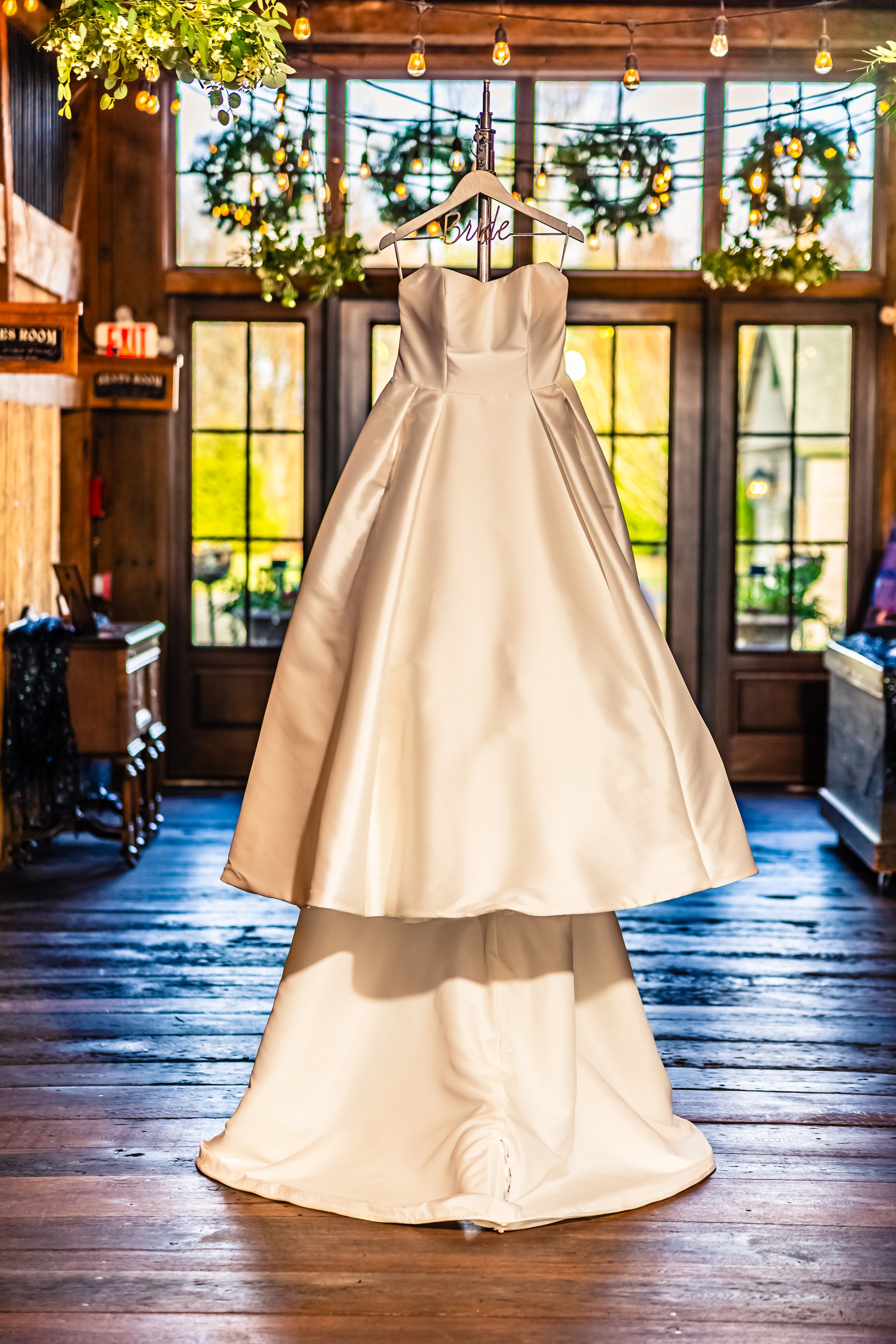 A white wedding dress hangs on a wooden hanger with a 'bride' sign in a rustic indoor setting with wooden walls, windows, green wreaths, and string lights.