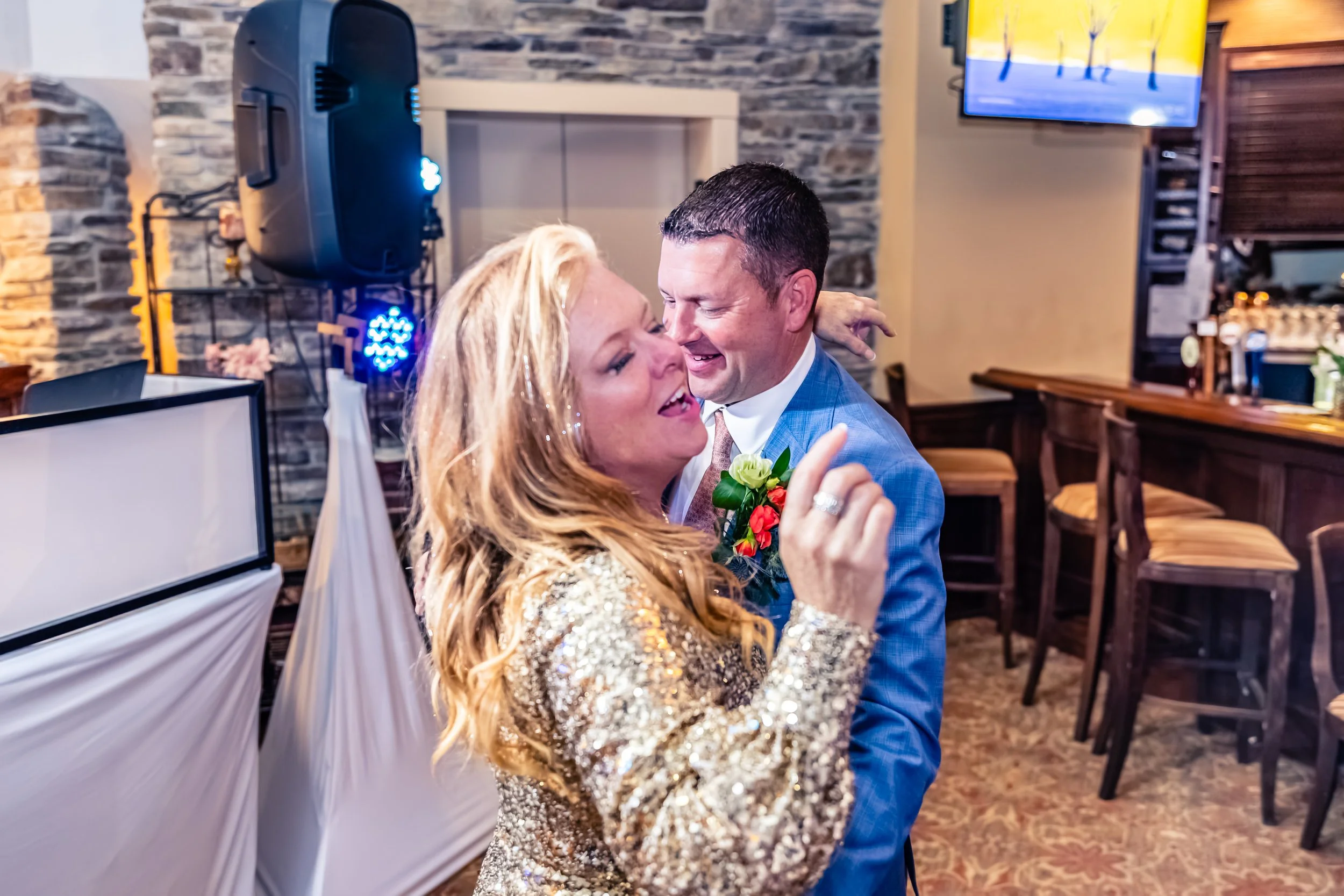 A man and a woman dancing closely together at a celebration or wedding reception, with the woman wearing a sparkly gold dress and the man in a blue suit with a boutonniere.