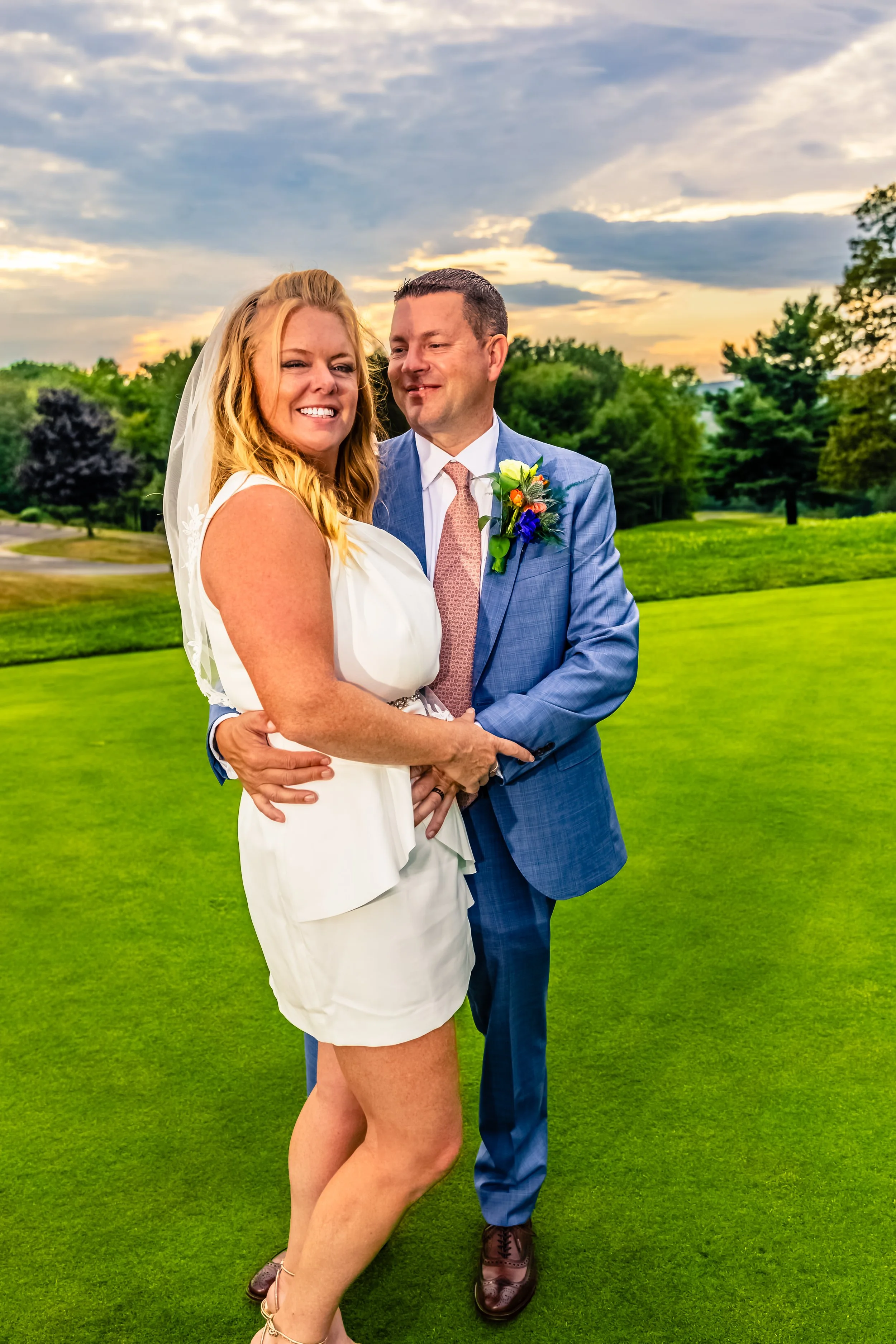 A smiling woman in a white dress and a man in a blue suit holding hands outdoors on a green field at sunset with clouds in the sky.