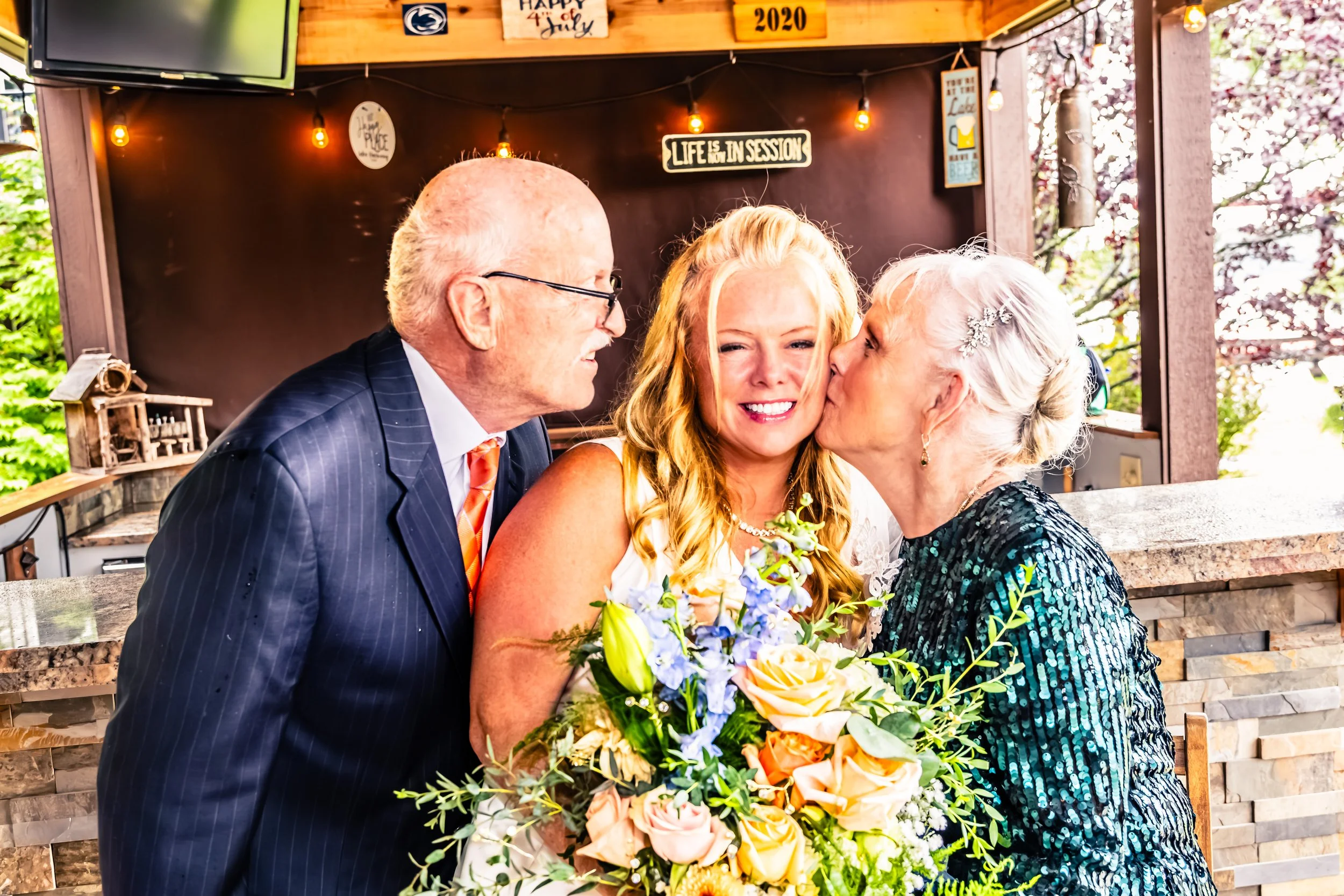 An elderly man and woman kiss a young woman with red hair and a large bouquet of flowers on her wedding day in a decorated outdoor setting.