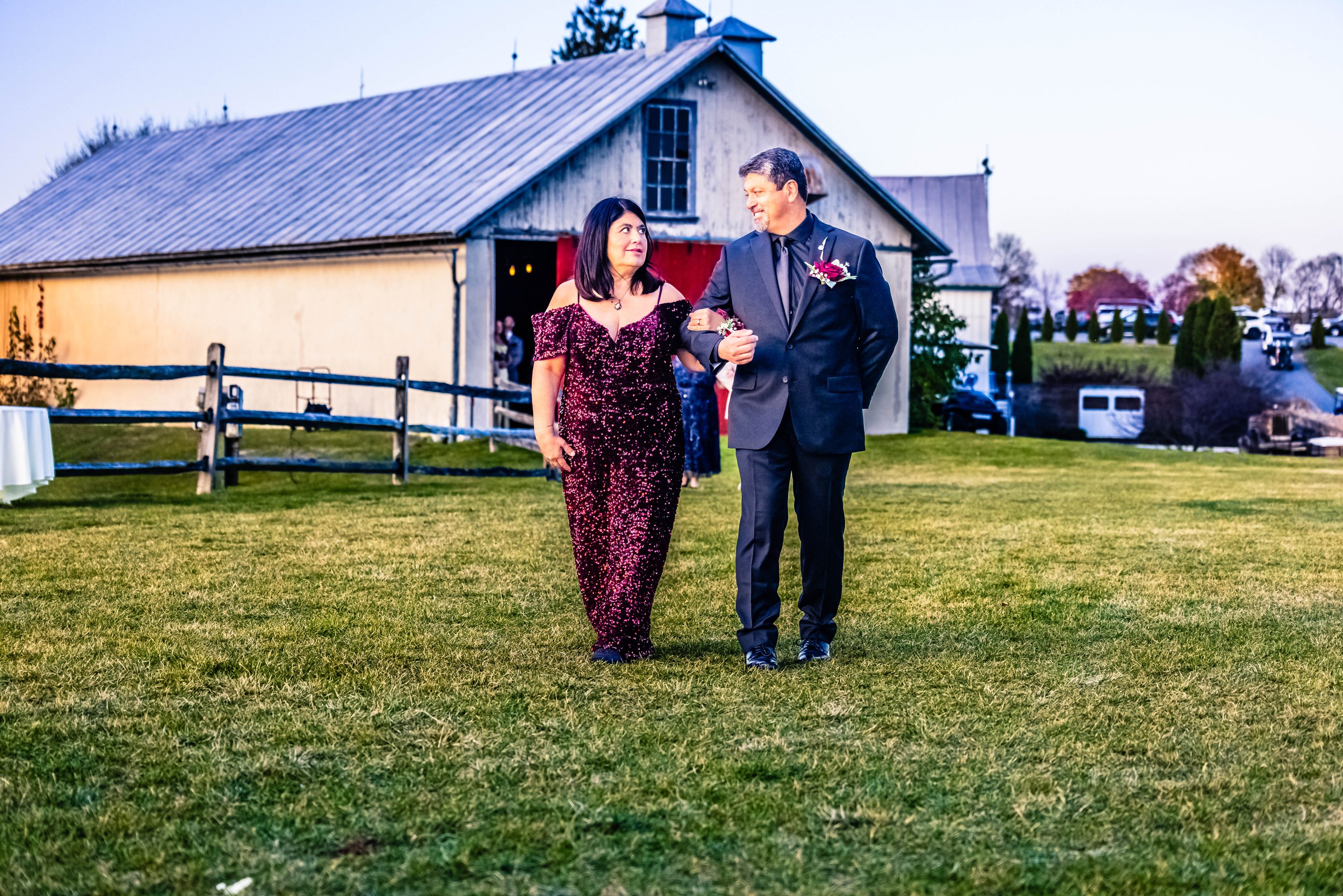 A woman and man walking arm in arm on a grassy field during a wedding celebration, with a barn and trees in the background.