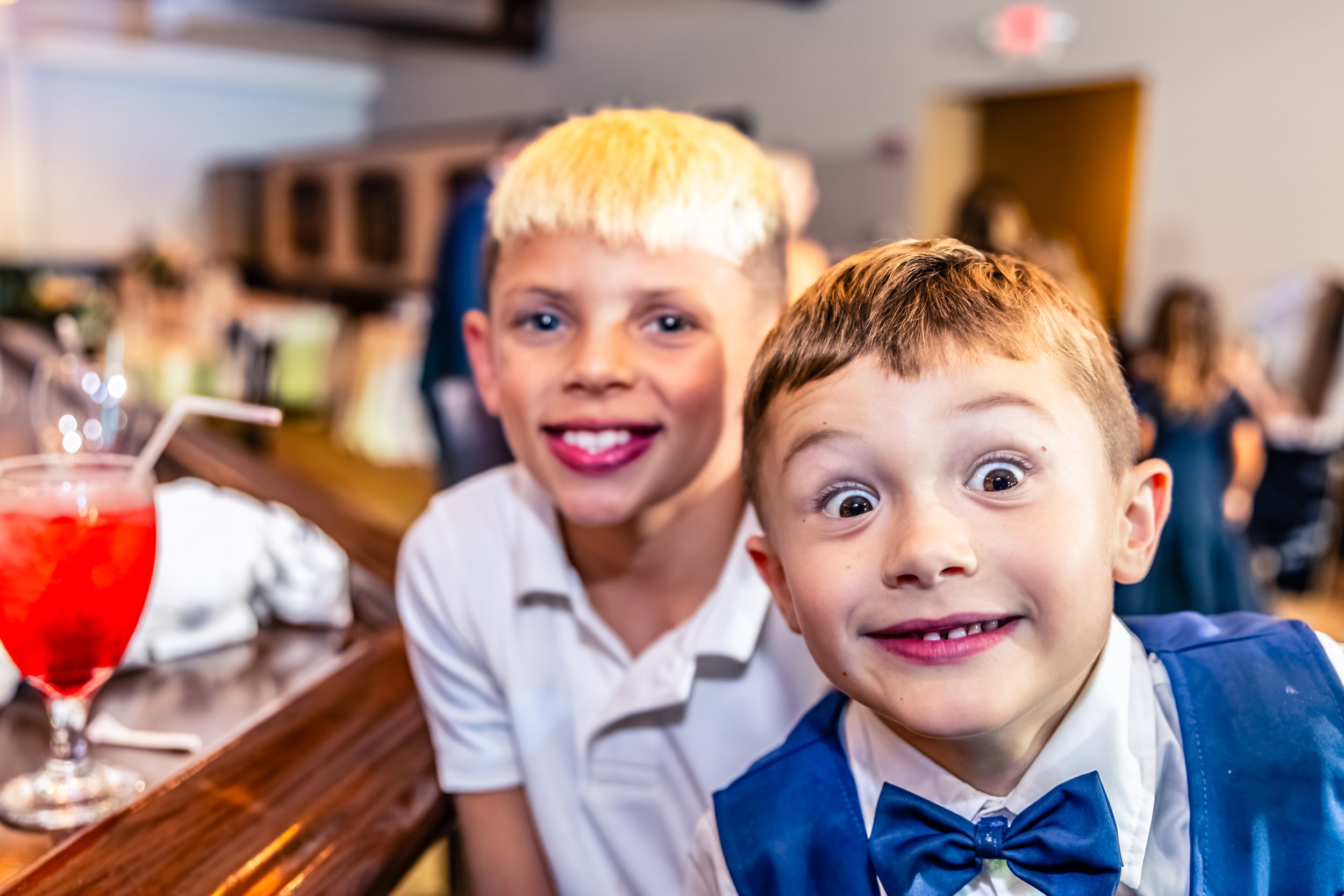 Two boys smiling at the camera at a social event. One wears a white shirt, the other a blue vest and bow tie. There is a red drink with a straw on the table.