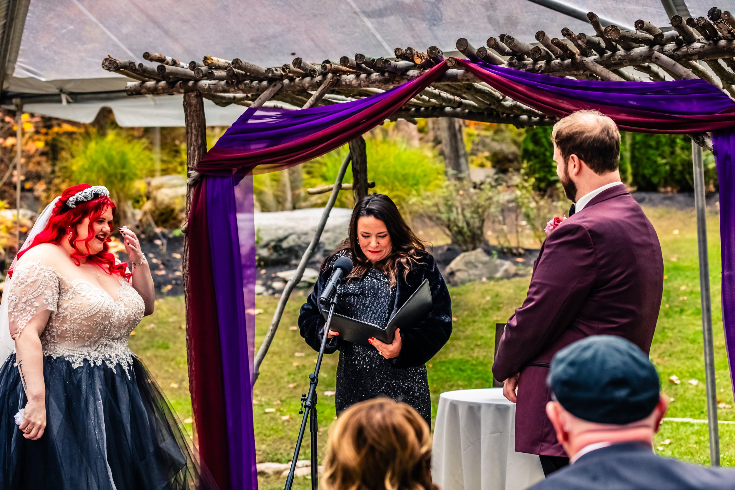 Bride with red hair in a wedding gown and a dark tulle skirt, standing under an outdoor wedding arch decorated with purple and pink fabric, smiling and crying, while the officiant reads vows at an outdoor ceremony, with groom in a maroon suit and gue