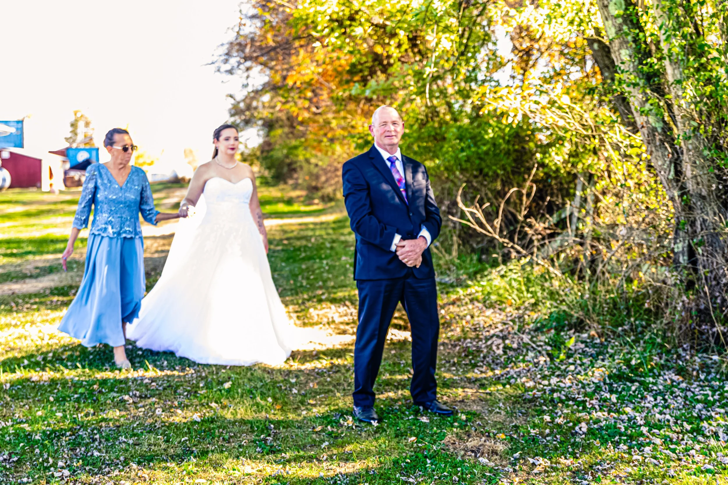 A man in a navy suit stands outdoors on a grassy area with trees, while a woman in a white wedding dress and another woman in a blue dress walk behind him. The background features colorful trees and buildings.