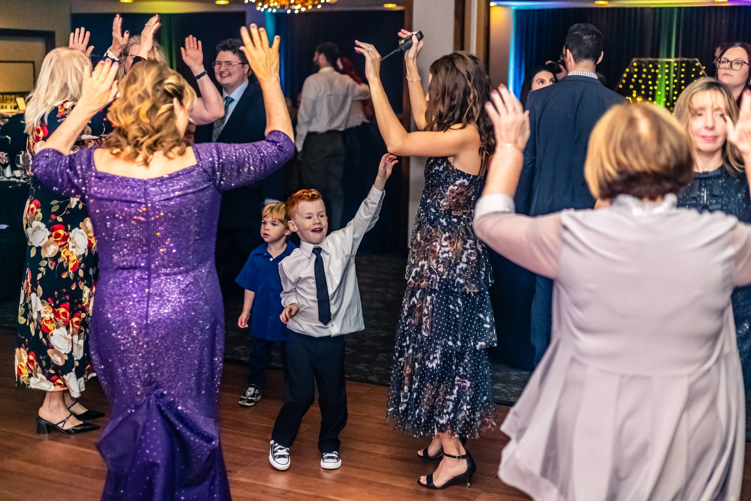 People dancing at a celebration or party, including children and adults, with colorful lighting in the background.