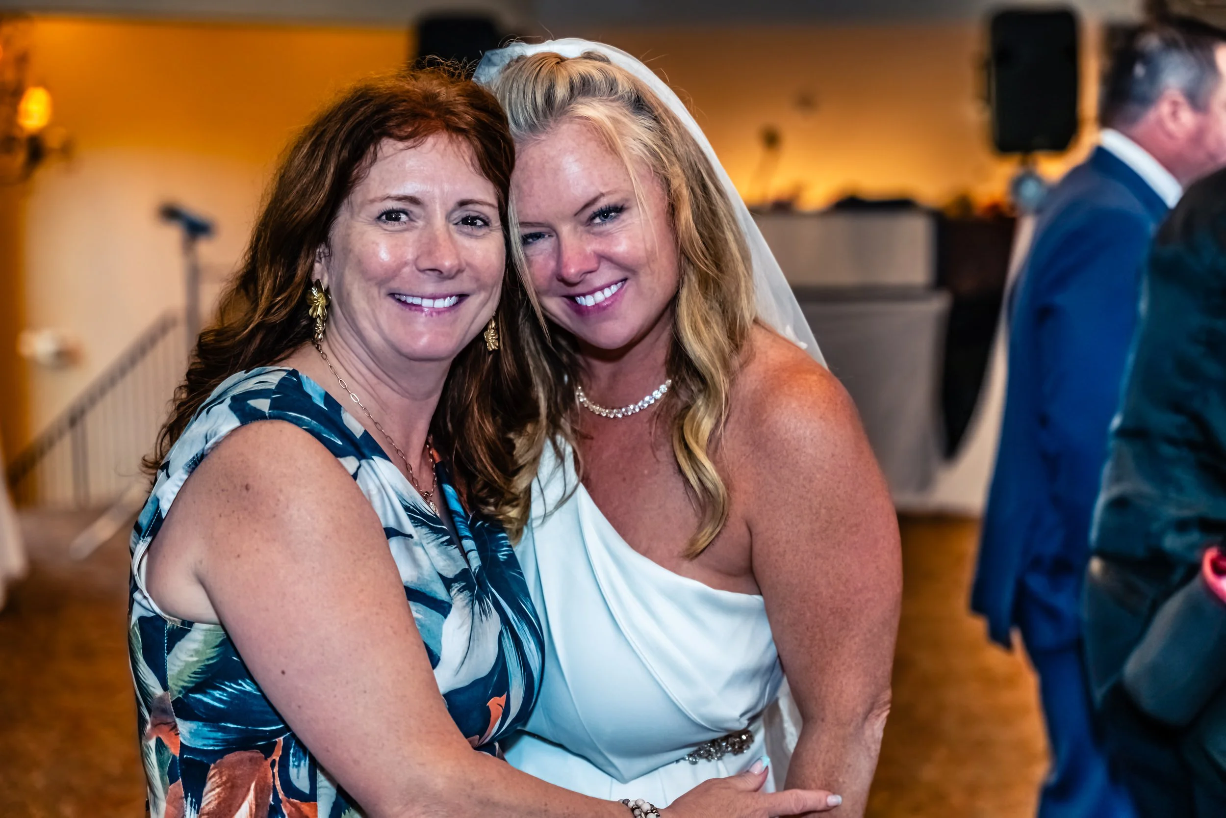 Two women smiling and posing for a photo at a wedding reception, with other guests in the background.