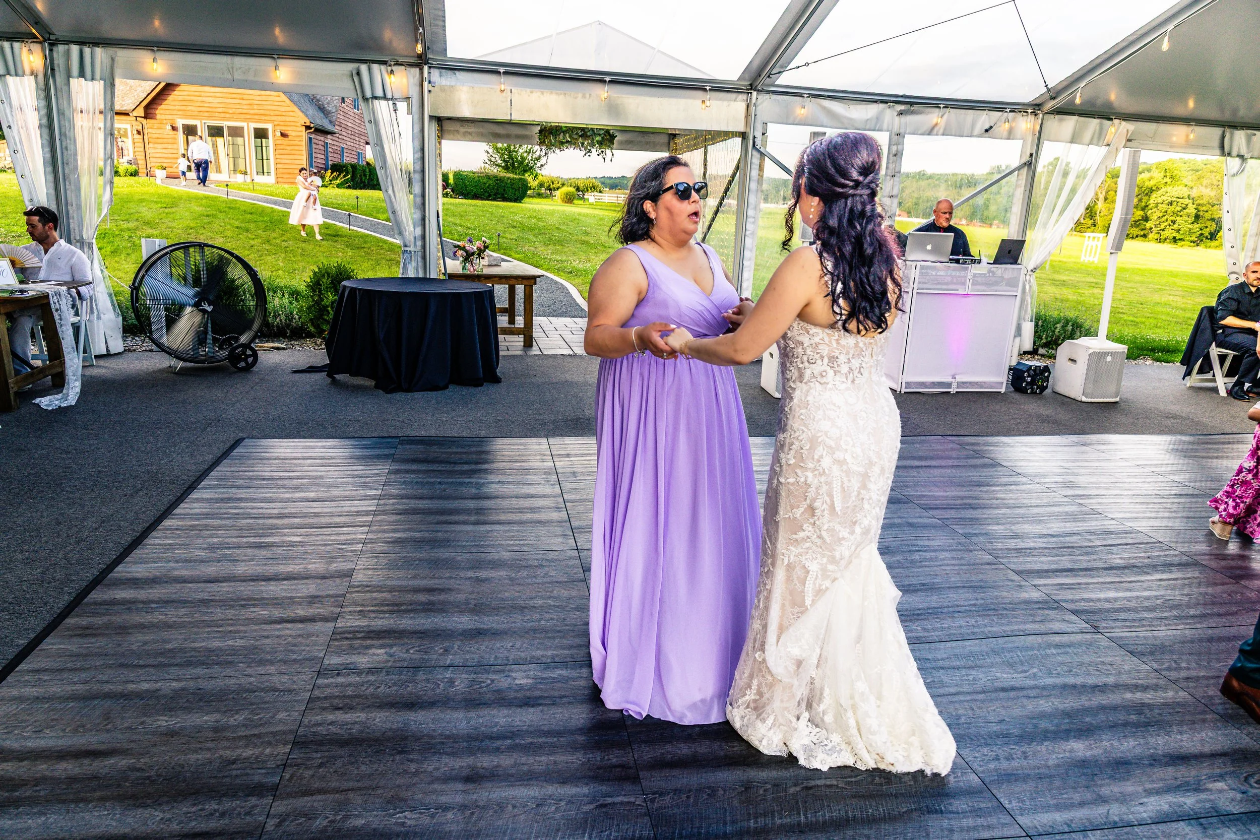Two women dancing at a wedding reception inside a tent, with a bride in a lace wedding dress and a guest in a purple gown. Other guests sit at tables and a DJ booth is visible in the background, with a scenic outdoor view through the tent openings.