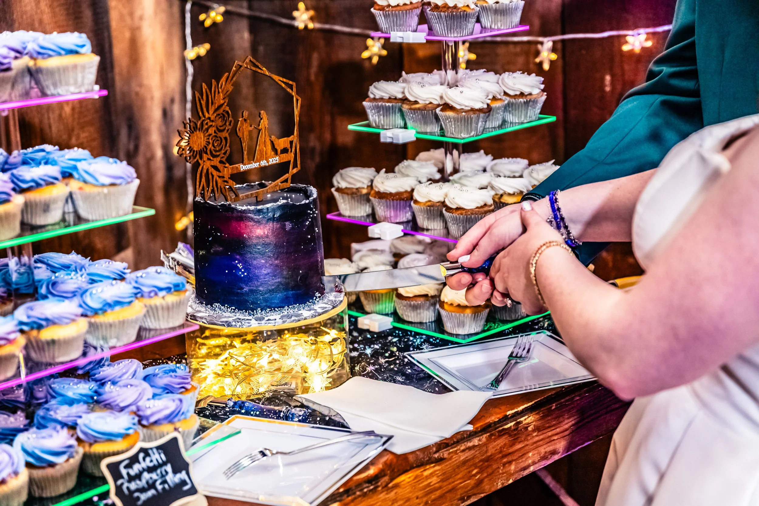 People cutting a galaxy-themed birthday cake at a celebration, with cupcakes decorated in blue and purple frosting visible on display.